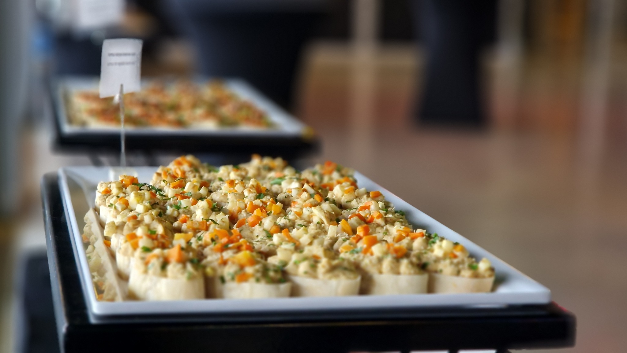 Taca z małymi kanapeczkami z pastą i posiekanymi warzywami (marchewka, ogórek, zioła) na białym prostokątnym talerzu, w tle rozmyte inne tace i elementy wyposażenia sali.