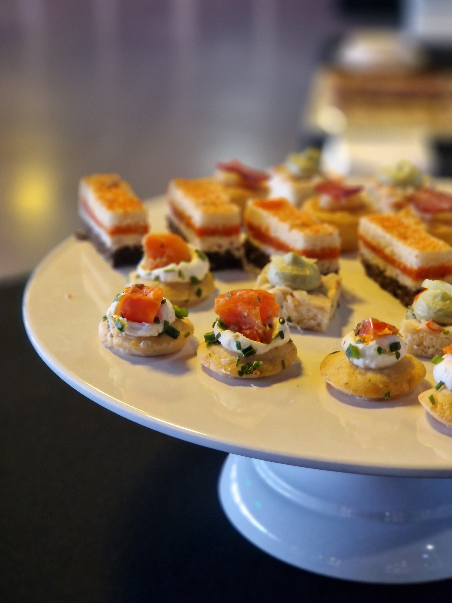 Różnorodne, apetyczne przekąski na białym, okrągłym paterze: krakersy z kremowym serem i łososiem, mini kanapeczki z pastami i wędlinami, ustawione w równych rzędach.