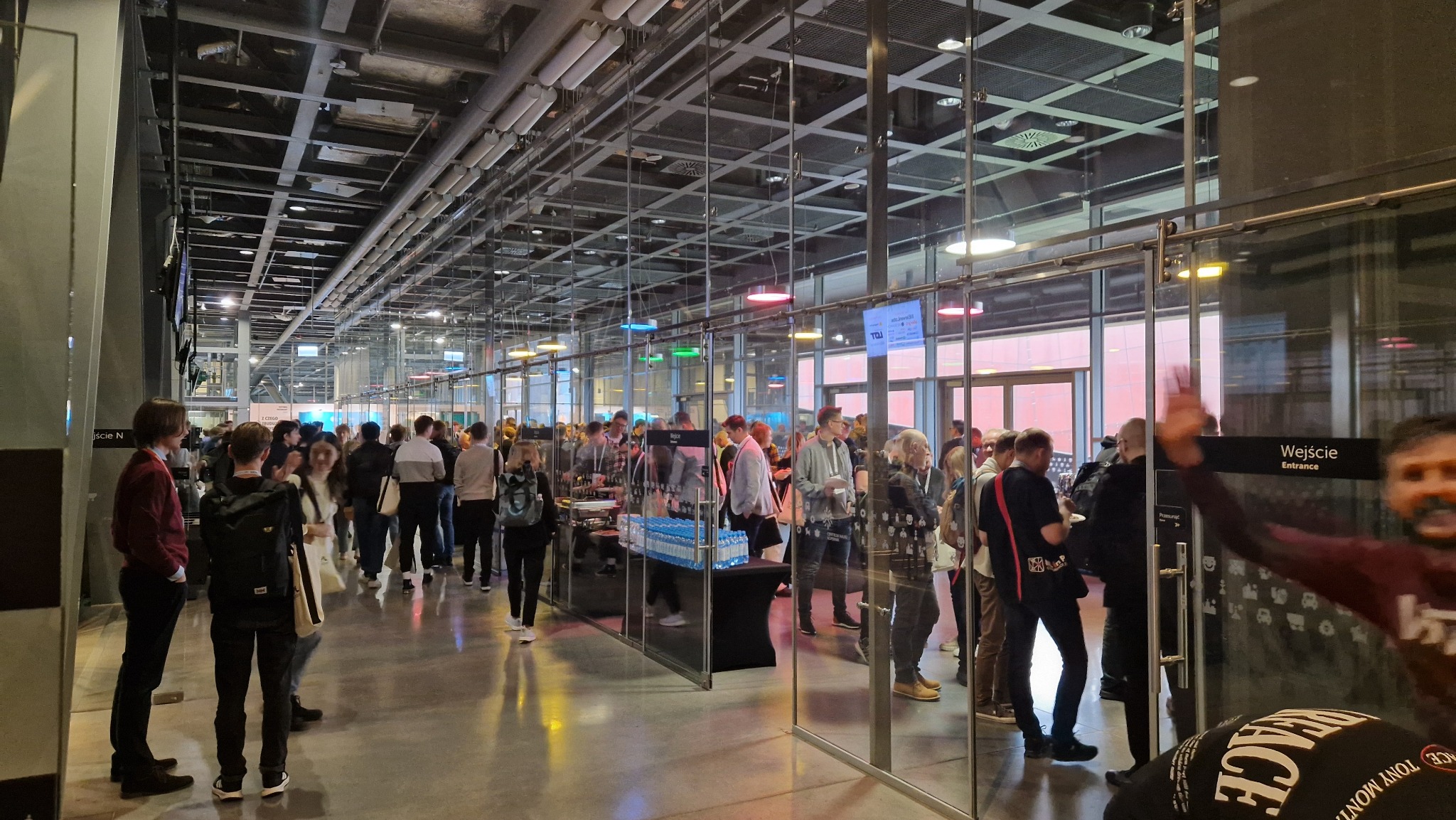 Wnętrze hali wystawienniczej z przeszklonymi ścianami, widoczny tłum ludzi, stoliki z napojami i przekąskami, surowy sufit z instalacjami.