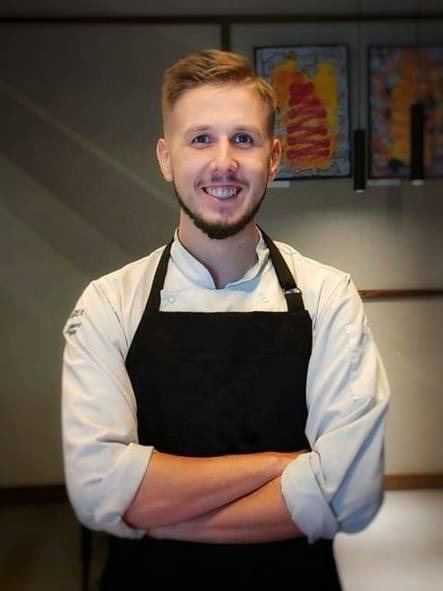 Uśmiechnięty kucharz w białej bluzie i czarnym fartuchu, z założonymi rękami na tle ściany z obrazami.