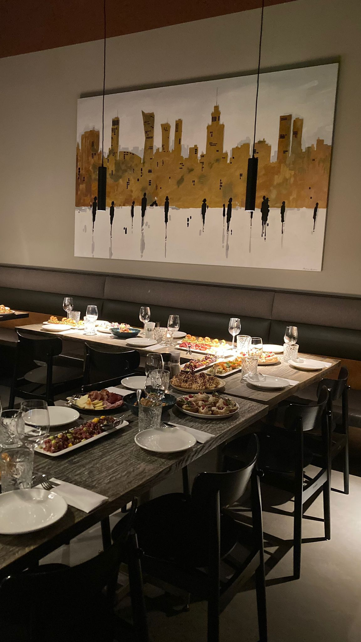 Ustawiony stół z przekąskami i szklankami na tle abstrakcyjnego obrazu przedstawiającego panoramę miasta, przygotowany do przyjęcia gości.