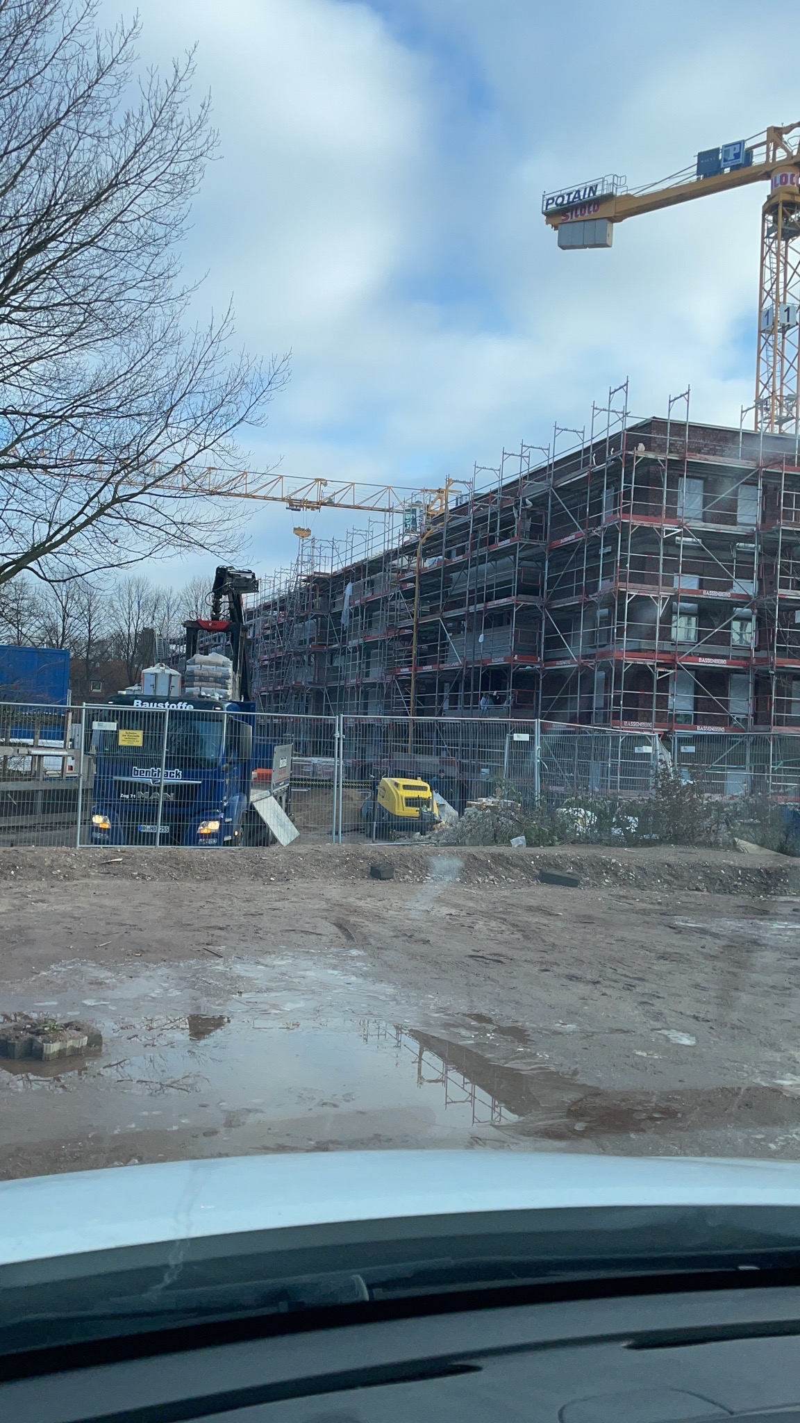 Budowa nowego budynku w Warszawie, widok przez przednią szybę samochodu, rusztowania na elewacji, dźwig budowlany i ciężarówka z materiałami budowlanymi.