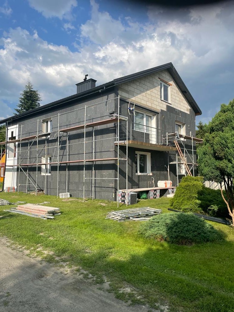 Dwukondygnacyjny dom w trakcie ocieplania, widoczne rusztowania i czarne płyty izolacyjne na elewacji, fragment niezabezpieczonej ściany z bloczków oraz rozłożone materiały budowlane na trawniku.