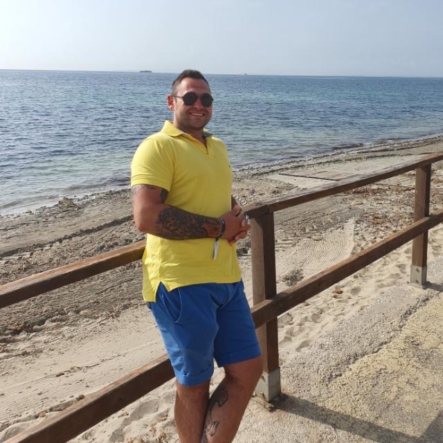 Mężczyzna w żółtej koszulce polo i niebieskich spodenkach stoi na plaży obok drewnianej barierki, z tatuażami na ramieniu i nodze, w tle morze i niebo.