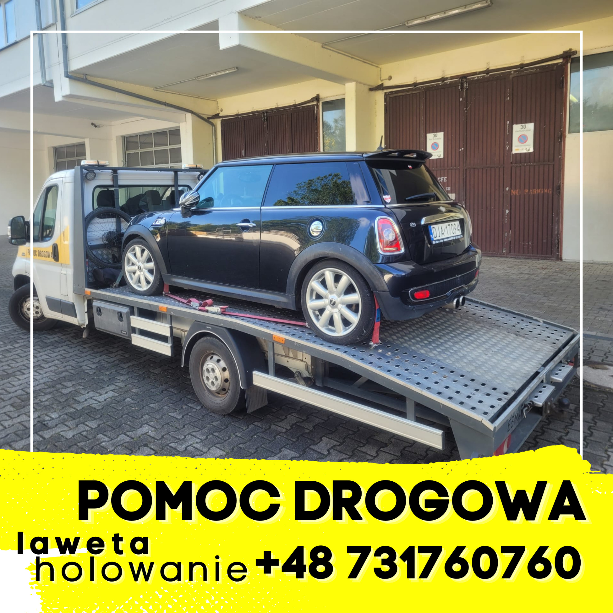 Czarny Mini Cooper na platformie lawety, zabezpieczony czerwonymi pasami, na tle budynku z brązowymi bramami garażowymi.