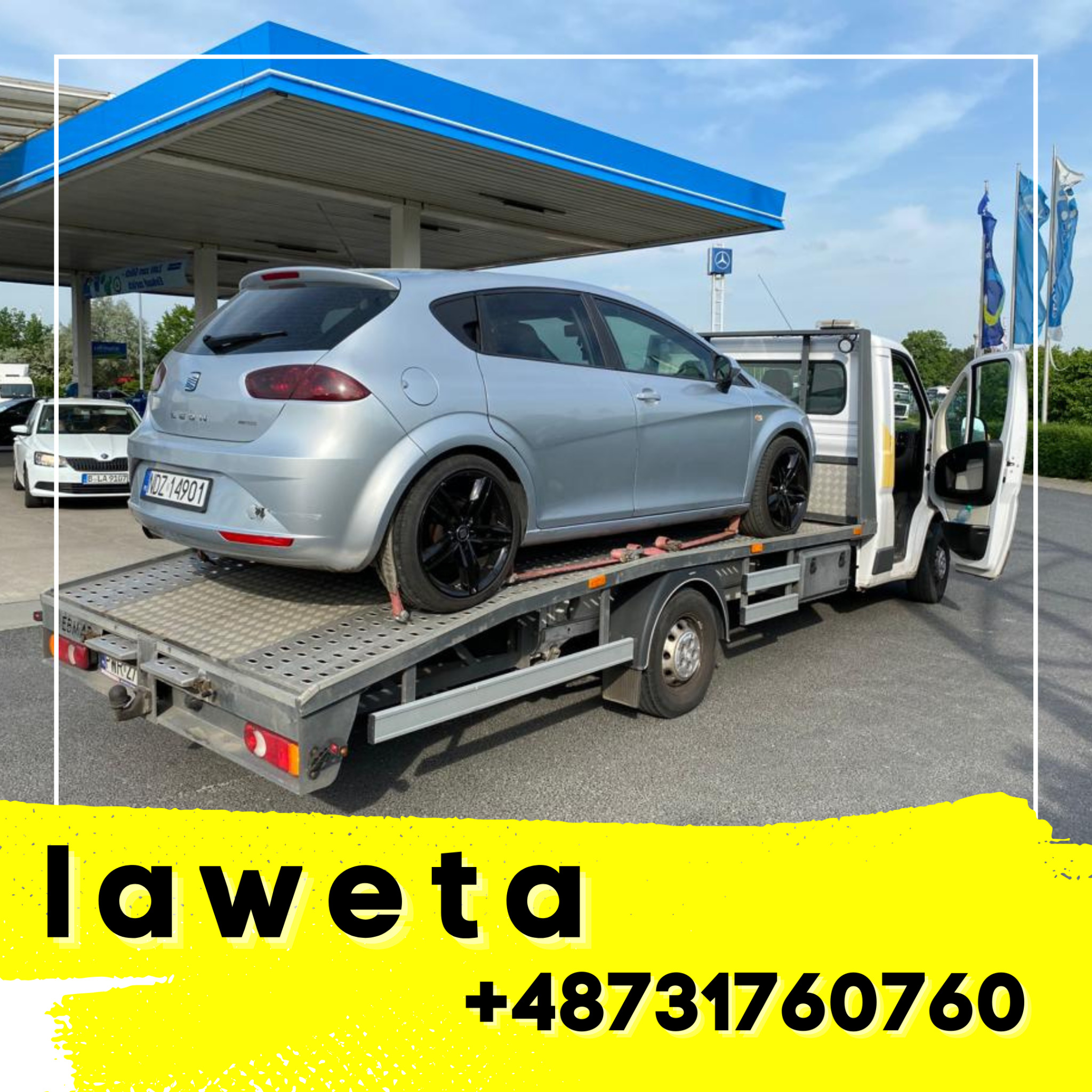 Laweta z szarym samochodem osobowym Seat Leon na platformie, zaparkowana przy stacji benzynowej z widocznymi flagami i otwartymi drzwiami kabiny kierowcy.