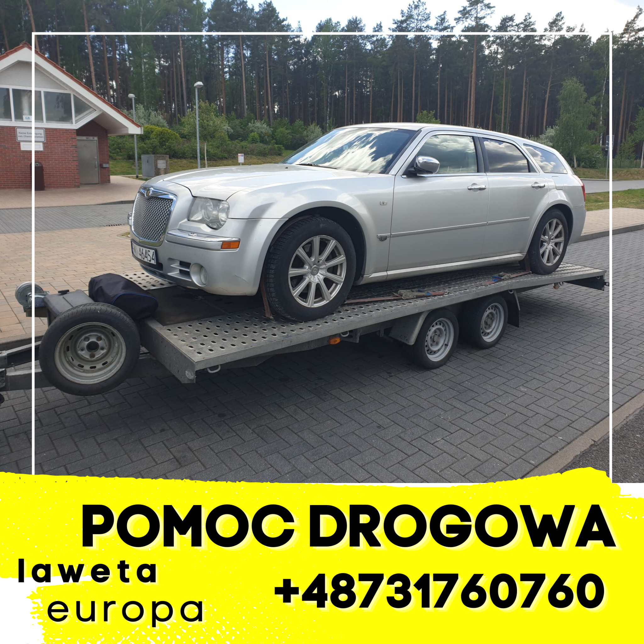 Srebrny Chrysler 300C kombi przewożony na dwuosiowej przyczepie lawetą, widoczny zapasowy opona i plandeka zabezpieczająca, w tle budynek toalety publicznej i las.