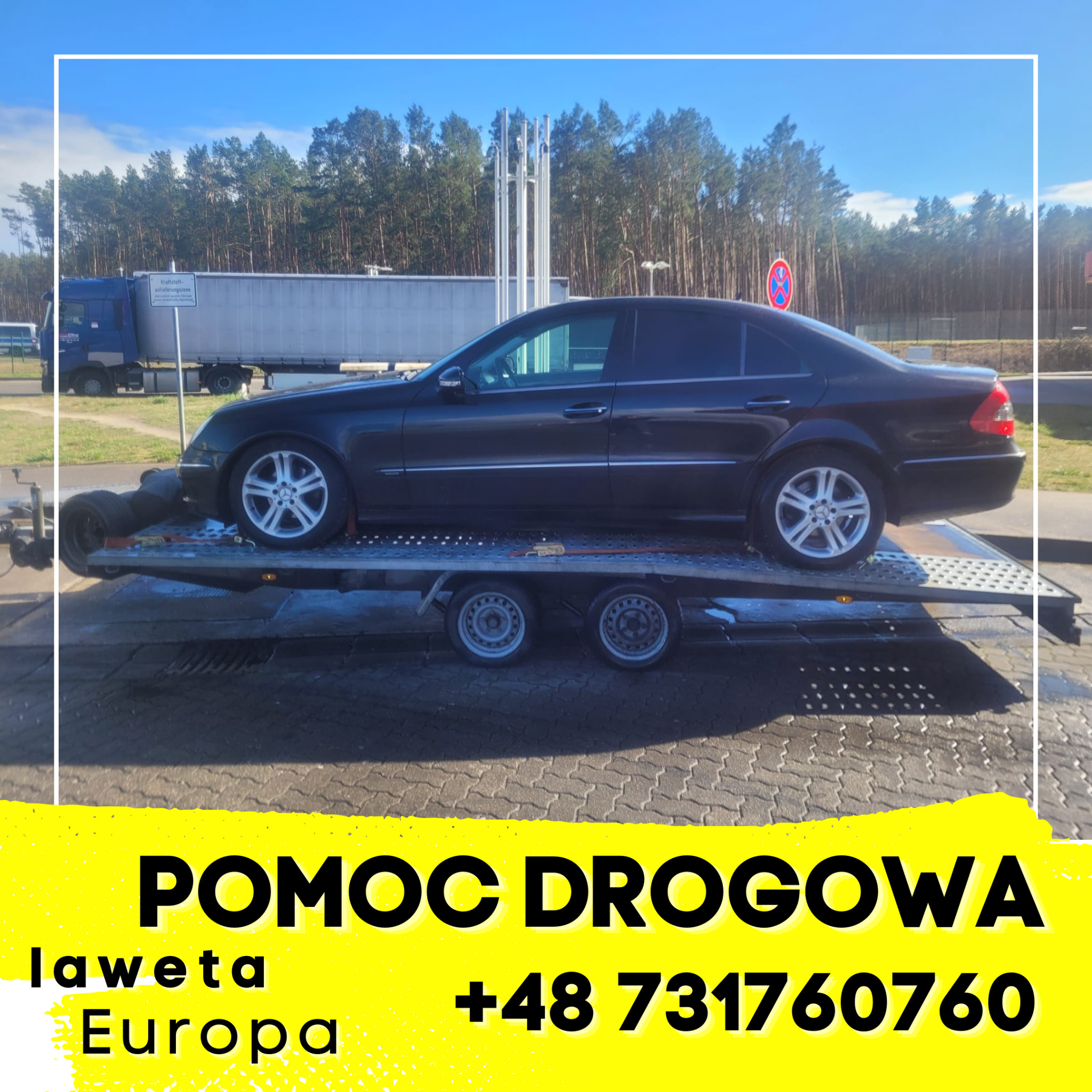 Czarny samochód osobowy marki Mercedes na platformie lawety, w tle widoczny las i budynki przemysłowe.