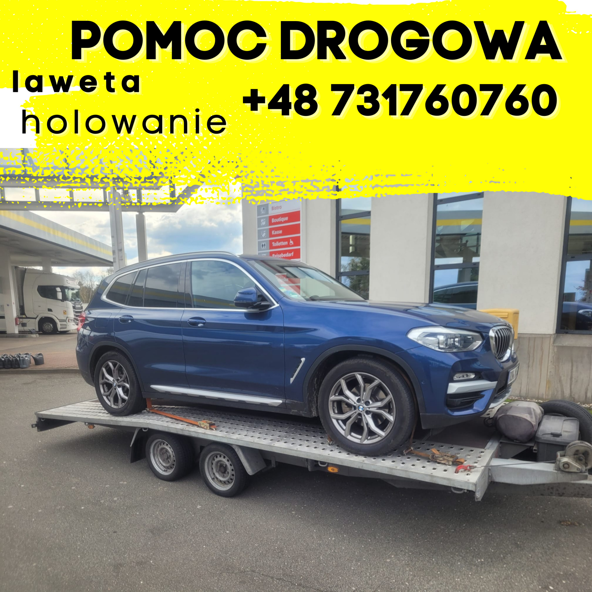 Niebieski samochód BMW X3 zabezpieczony pasami na lawecie, widok z boku, w tle budynek stacji benzynowej i zaparkowany biały samochód ciężarowy.