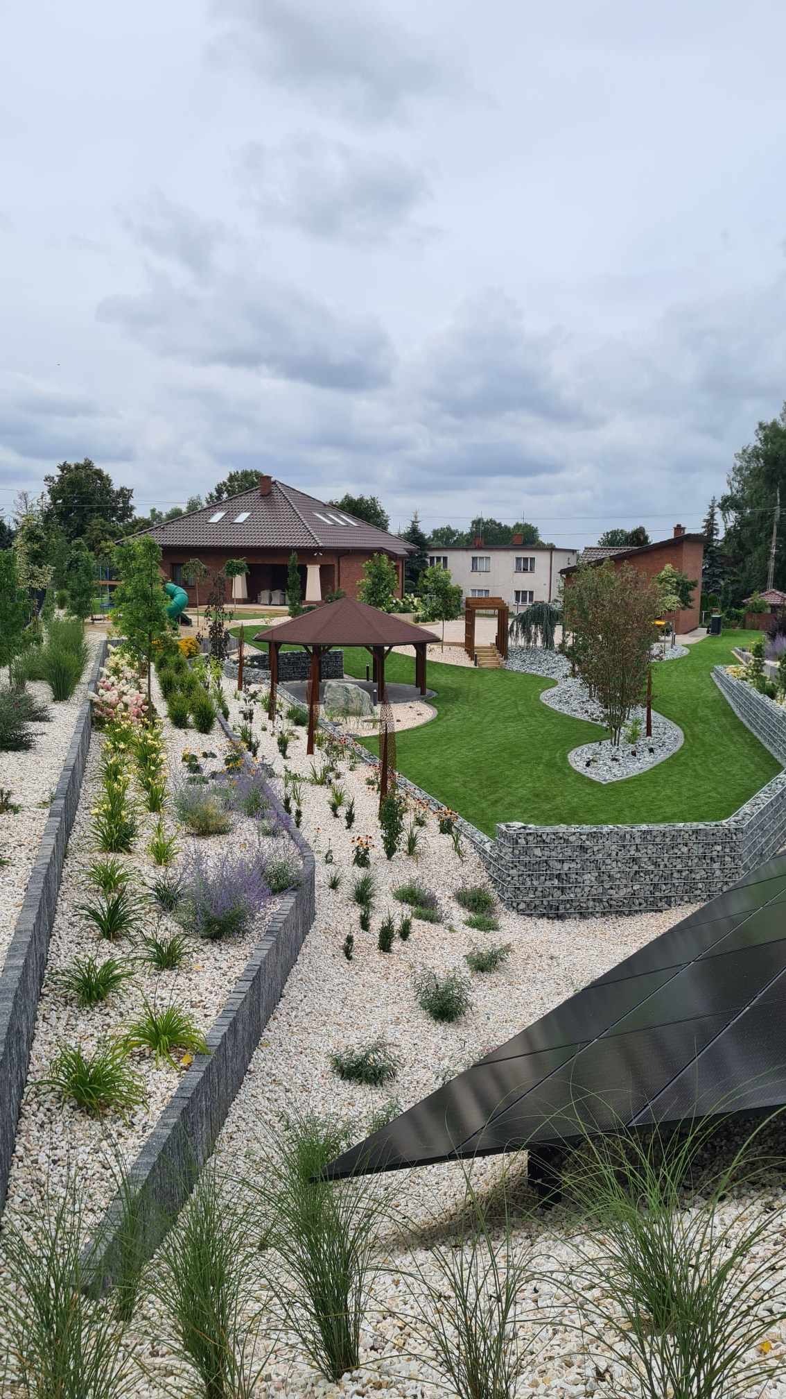 Widok na starannie zaprojektowany ogród z altaną, trawnikiem, rabatami z kamyków i roślin oraz gabionami, na tle domu jednorodzinnego w pochmurny dzień.