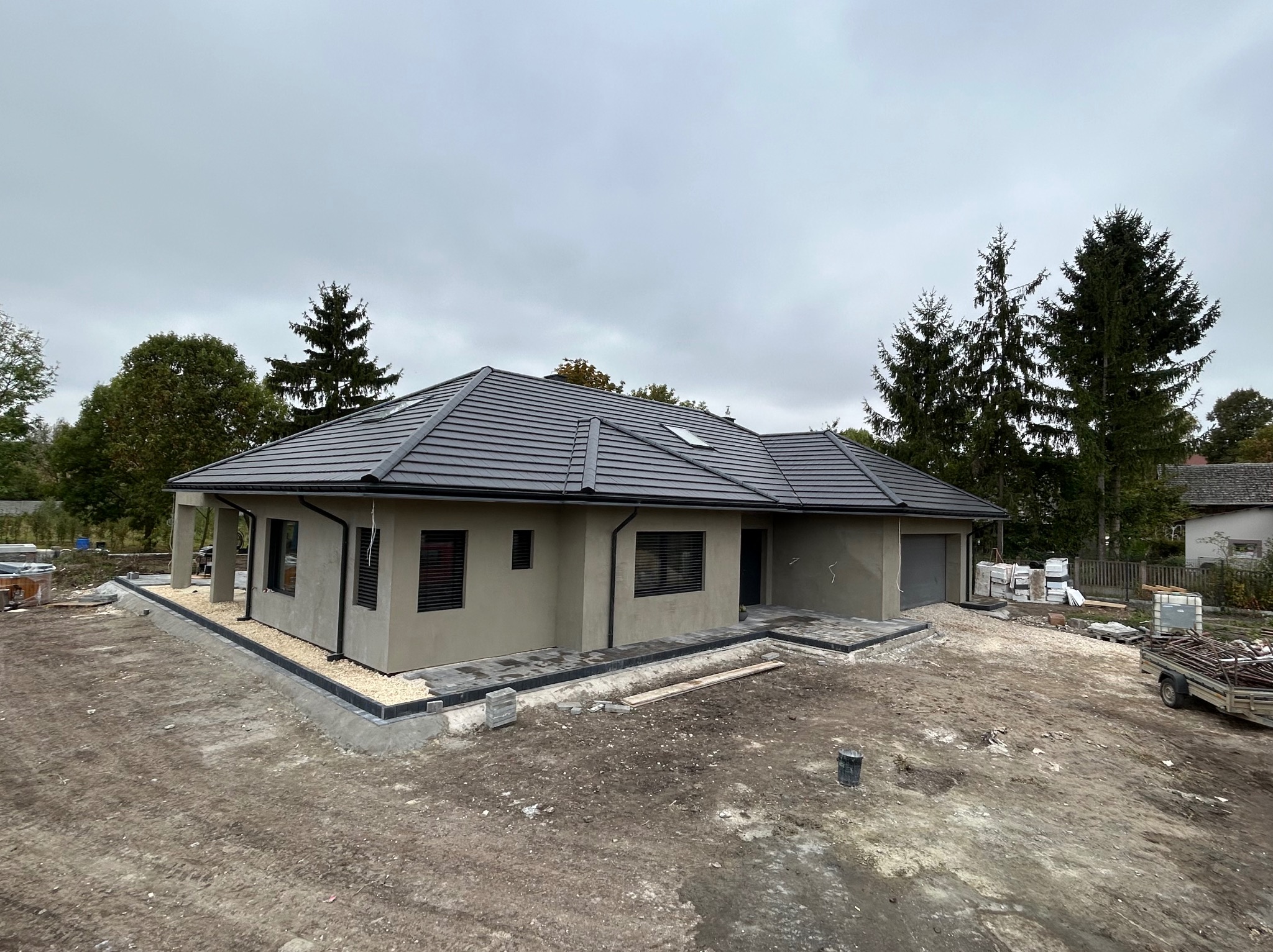 Parterowy dom z szarą elewacją i ciemną dachówką, widoczny z boku, z elementami wykończeniowymi wokół budynku. W tle drzewa i pochmurne niebo.