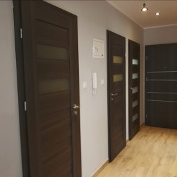 Profesjonalne wykanczanie wnętrz - Korytarz z trzema zamontowanymi drzwiami wewnętrznymi w odcieniach brązu, widoczne panele podłogowe, domofon i skrzynka elektryczna na ścianie.