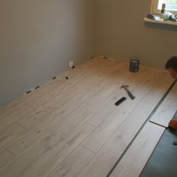 Profesjonalne wykanczanie wnętrz - Układanie paneli podłogowych w rogu pokoju, widoczne szczeliny dylatacyjne z klinami, młotek i puszka farby na parapecie okna.