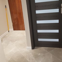 Profesjonalne wykanczanie wnętrz - Zamknięte, nowoczesne drzwi wewnętrzne z ciemnego drewna z poziomymi, jasnymi paskami, widoczne na tle szarej, marmurkowej podłogi z płytek i fragmentu brązowych drzwi wejściowych.