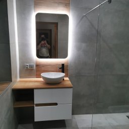 Profesjonalne wykanczanie wnętrz - Nowoczesna łazienka z szarymi płytkami, podświetlanym lustrem, białą umywalką nablatową i drewnianym blatem, widoczny fragment kabiny prysznicowej.