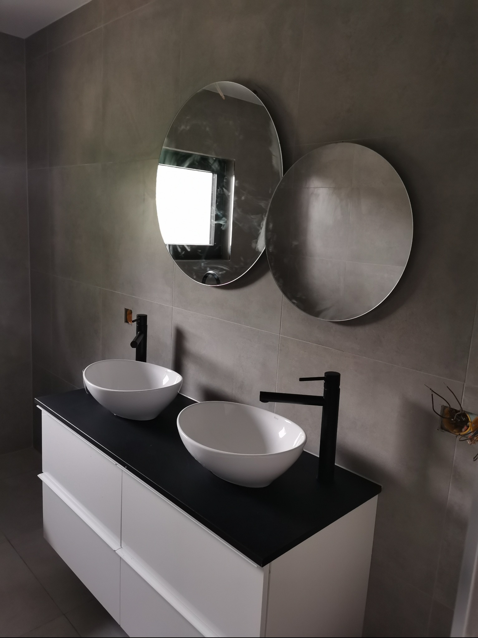 Nowoczesna łazienka z dwoma owalnymi umywalkami nablatowymi, czarnymi bateriami i dwoma okrągłymi lustrami na szarych płytkach, z widocznymi przewodami elektrycznymi w ścianie po prawej stronie.