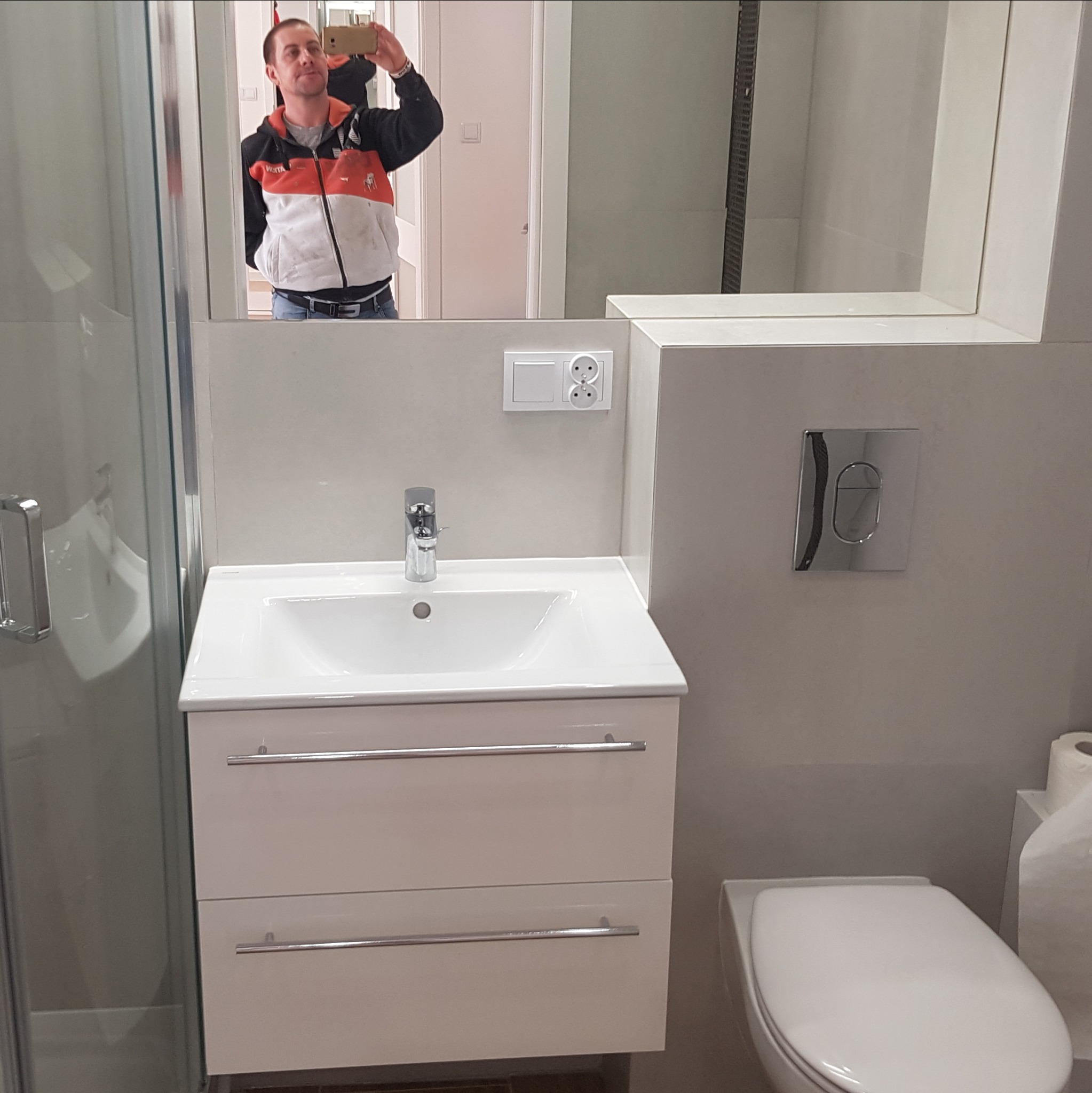 Nowoczesna łazienka z białą umywalką nablatową na szafce z dwoma szufladami, toaleta podwieszana z chromowanym przyciskiem spłukiwania i kabina prysznicowa, odbicie fotografa w lustrze.