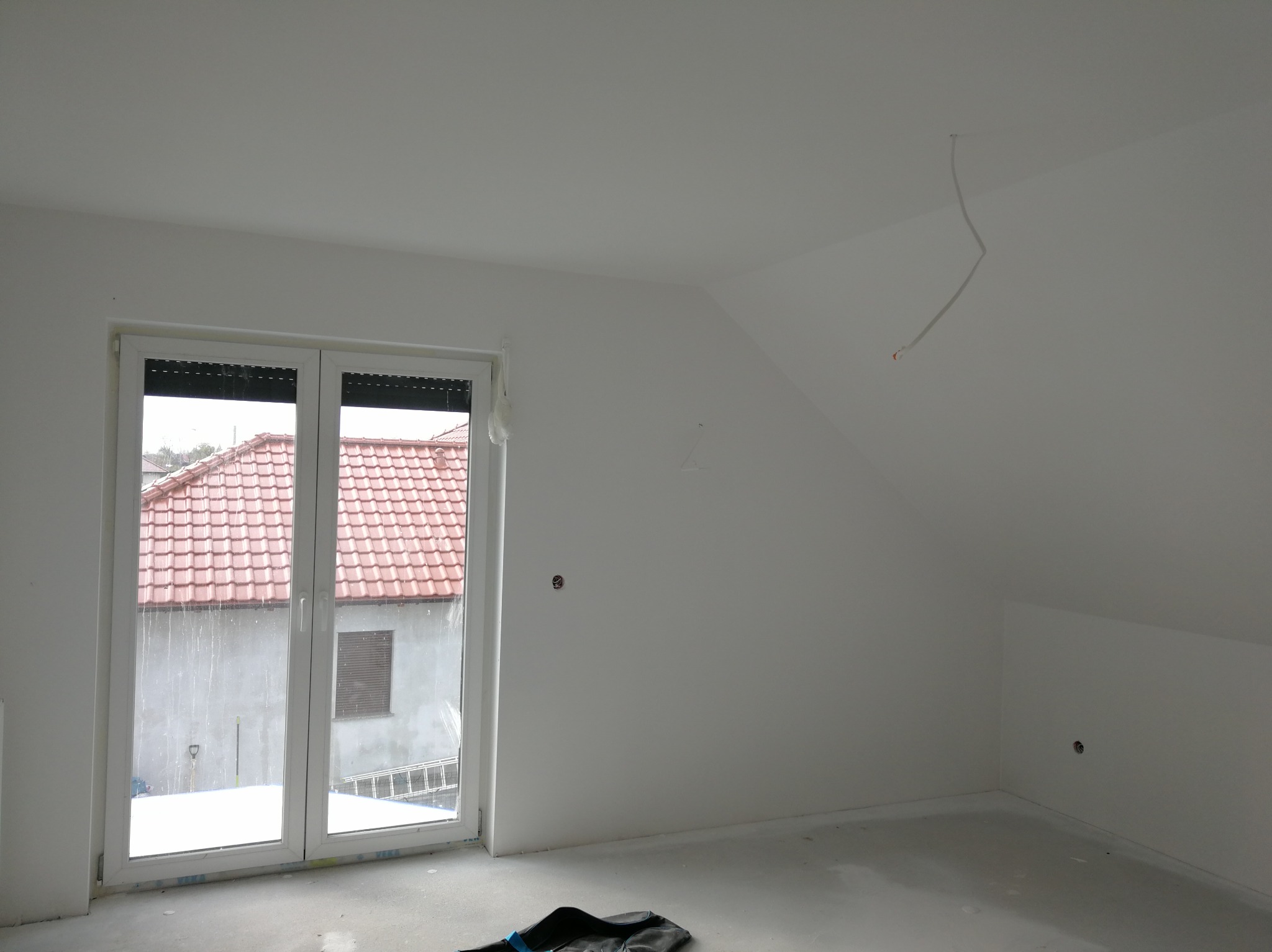 Poddasze w trakcie malowania na biało, widoczne gładkie ściany, okno z roletami i wystające kable ze ściany