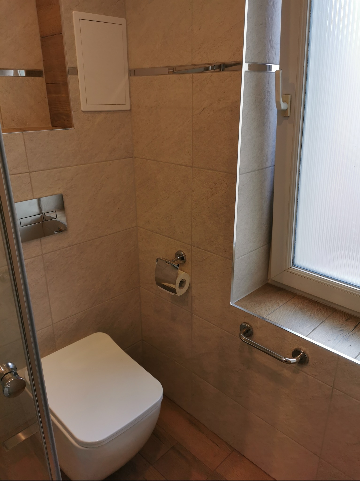 Nowoczesna toaleta podwieszana z chromowanym przyciskiem spłukującym i uchwytem dla niepełnosprawnych, na tle beżowych płytek imitujących kamień i okna z mleczną szybą.