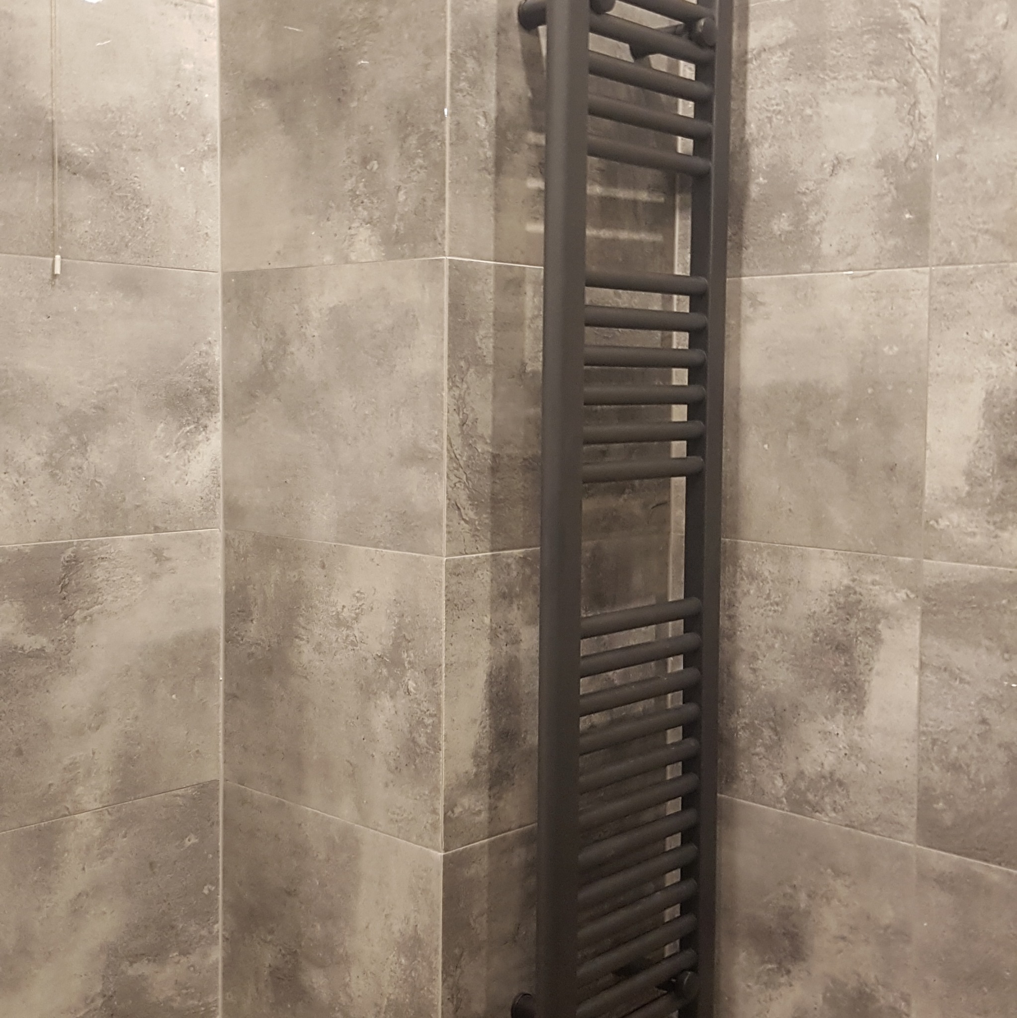 Szara, kwadratowa glazura ścienna z zamontowanym, pionowym, czarnym grzejnikiem drabinkowym w łazience.