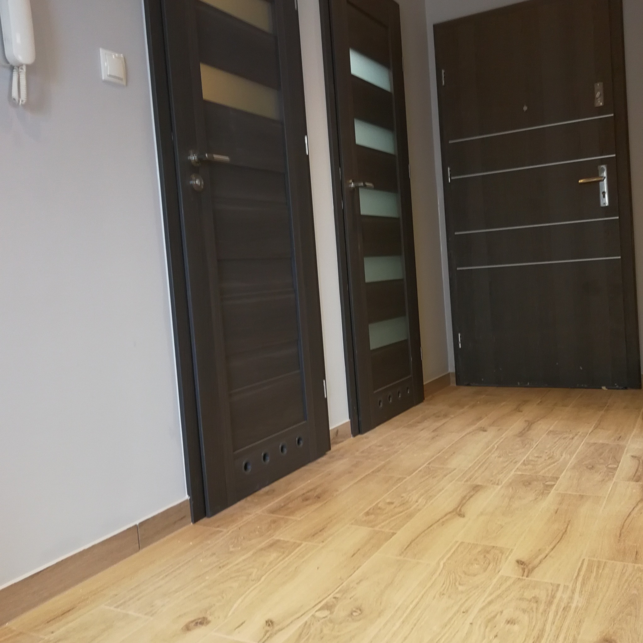 Trzy zamontowane, ciemnobrązowe drzwi wewnętrzne o różnym wzornictwie, widoczne na tle jasnej ściany i podłogi z płytek imitujących drewno.