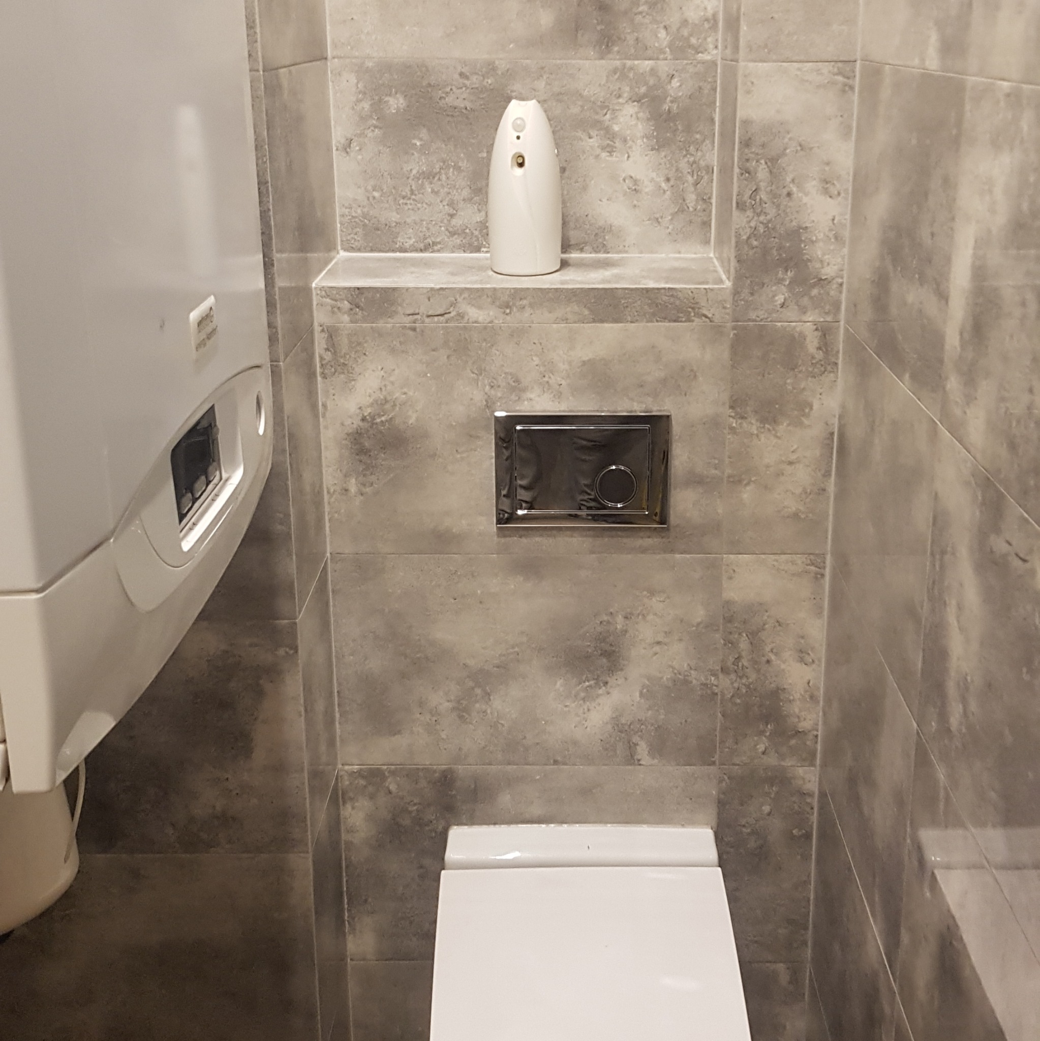 Szare, marmurkowe płytki w łazience, widoczny fragment toalety, chromowany przycisk spłuczki, biały automatyczny odświeżacz powietrza na półce i biały bojler.