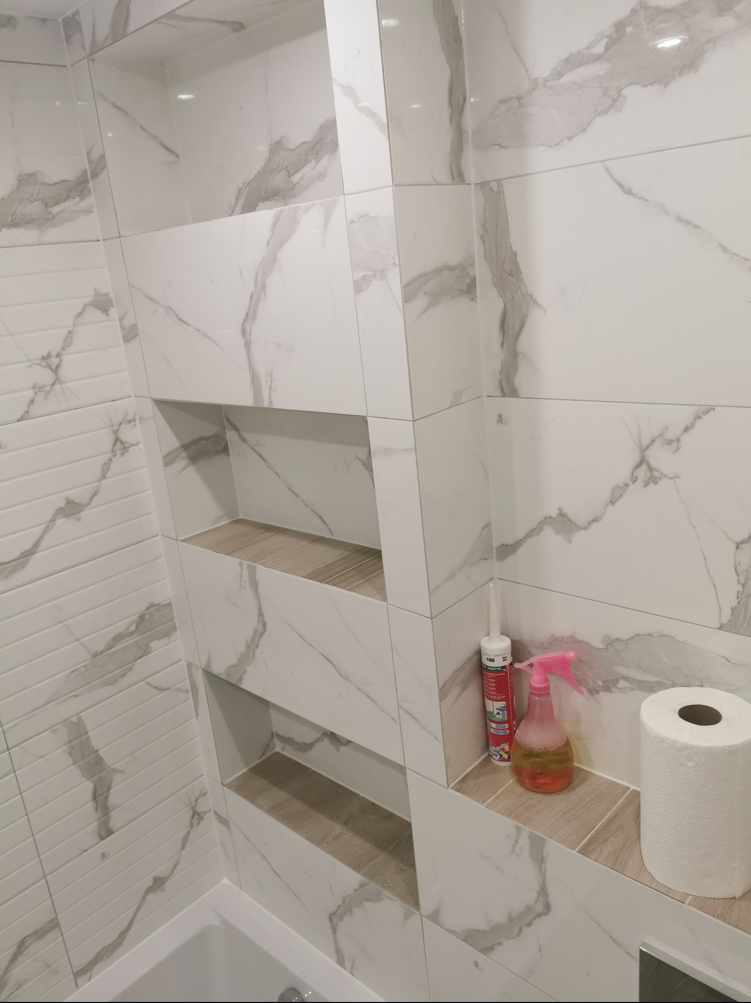 Ściana łazienki wyłożona białymi płytkami imitującymi marmur z wnękami wykończonymi drewnopodobnymi panelami. W jednej z wnęk biały silikon, różowy spryskiwacz i rolka papieru toaletowego.