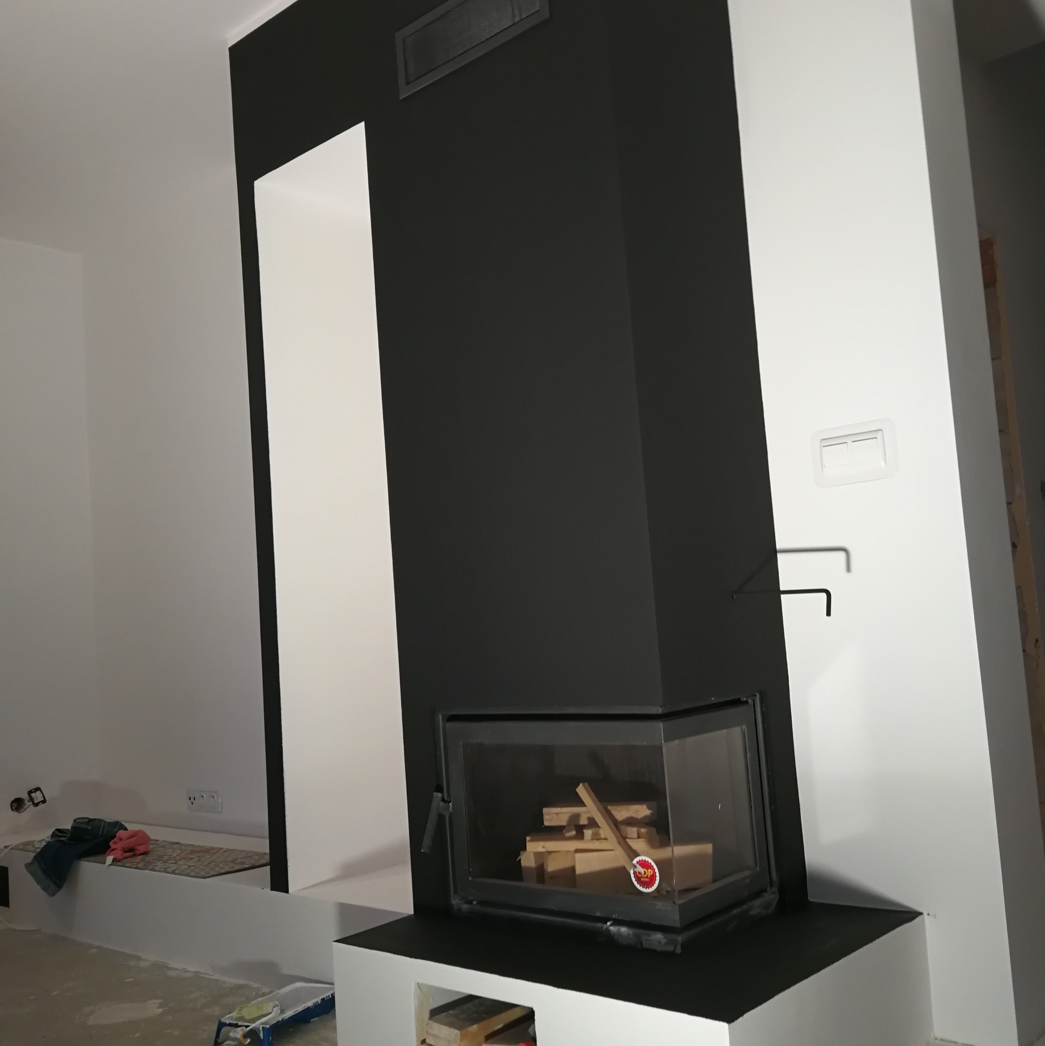Nowoczesny kominek z czarną obudową i wkładem szklanym, z ułożonym drewnem w środku, na tle białych ścian i czarnego pasa na ścianie, widoczne ślady prac wykończeniowych.