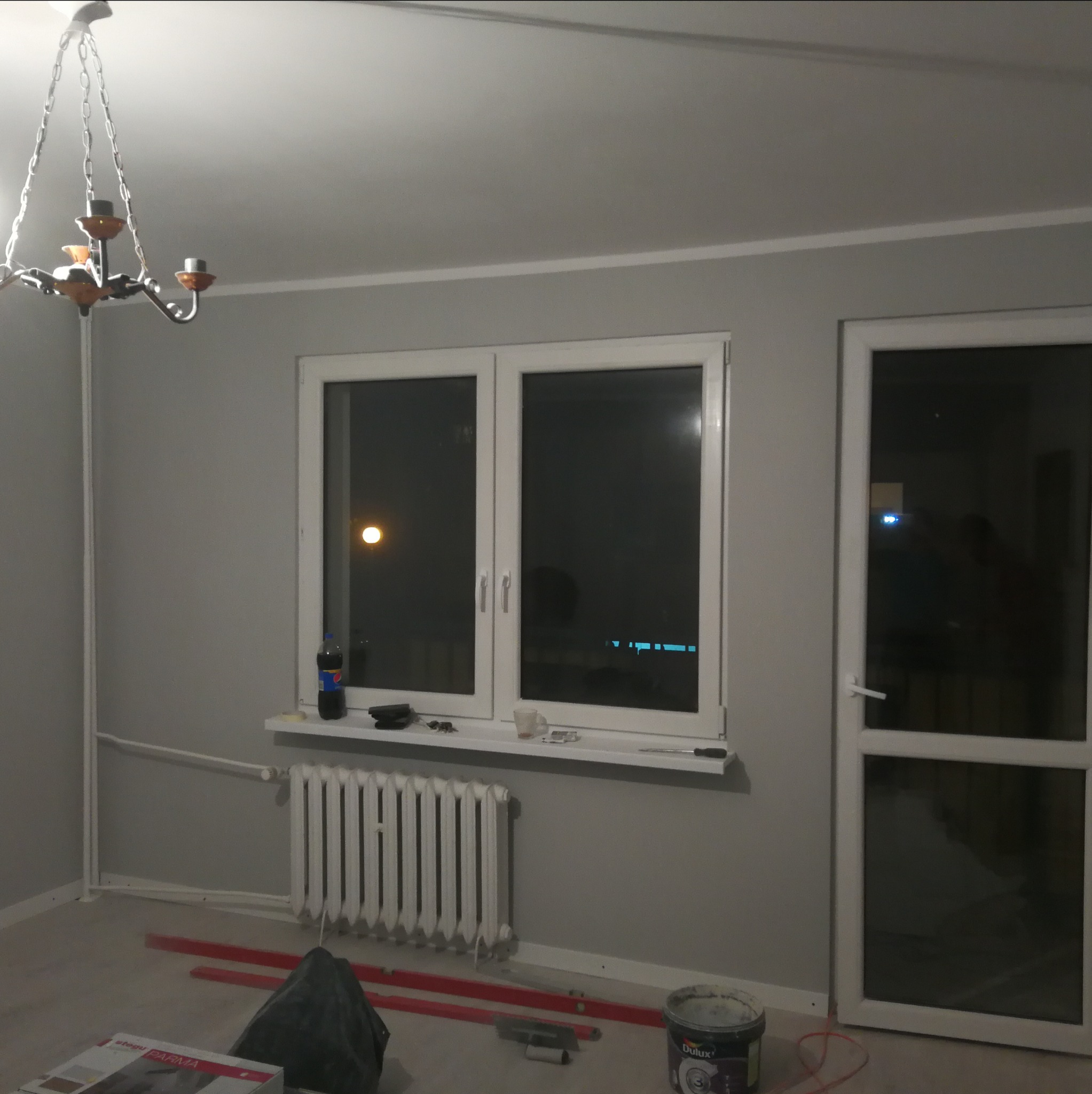 Pokój w trakcie remontu: szare ściany, białe okna, grzejnik, poziomice, wiadro z farbą Dulux, lampa wisząca i częściowo zakryte podłogi.