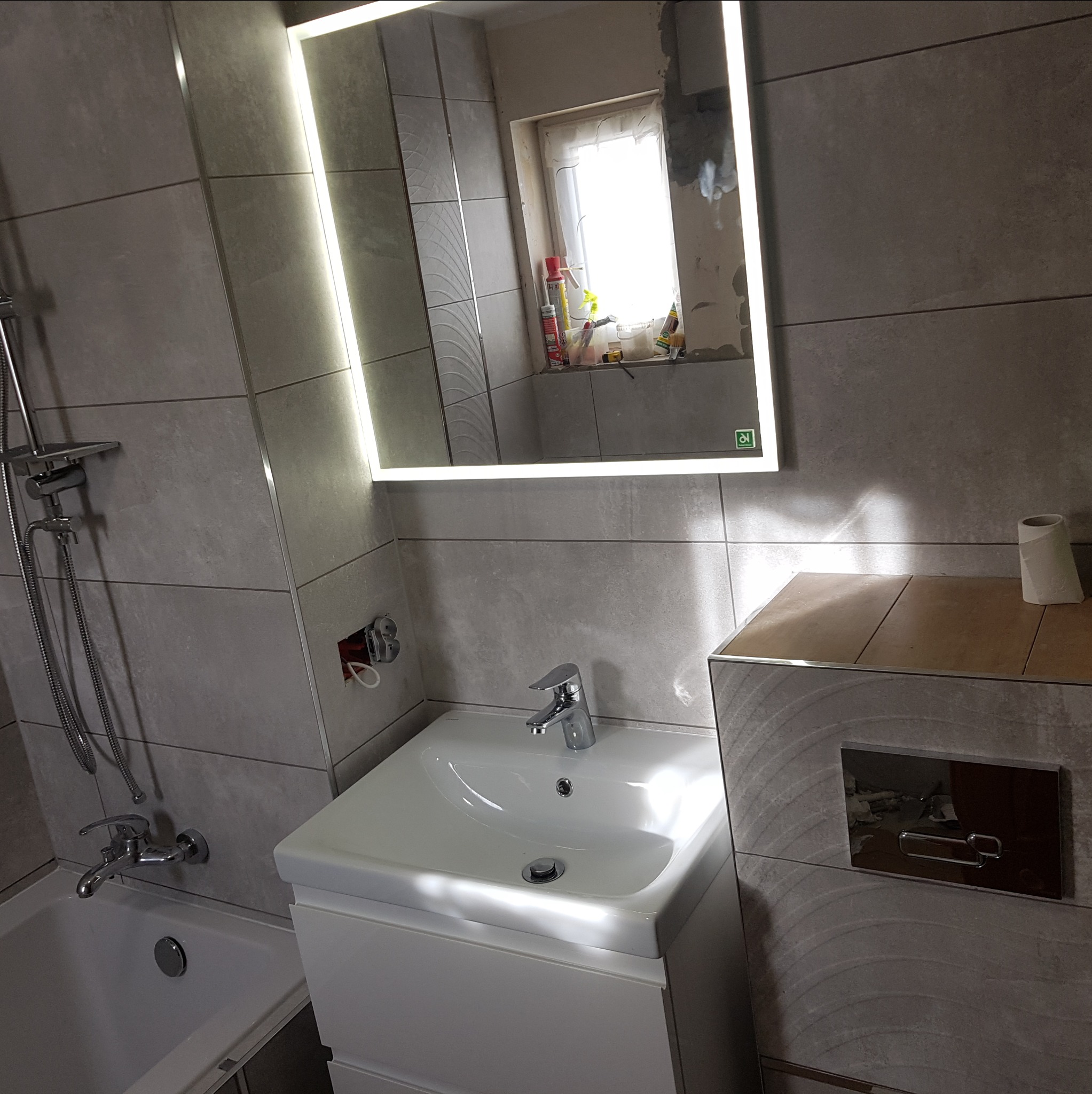 Wykończona łazienka z szarymi płytkami, podświetlanym lustrem i białą umywalką z szafką, widoczne niedokończone prace w odbiciu lustra.