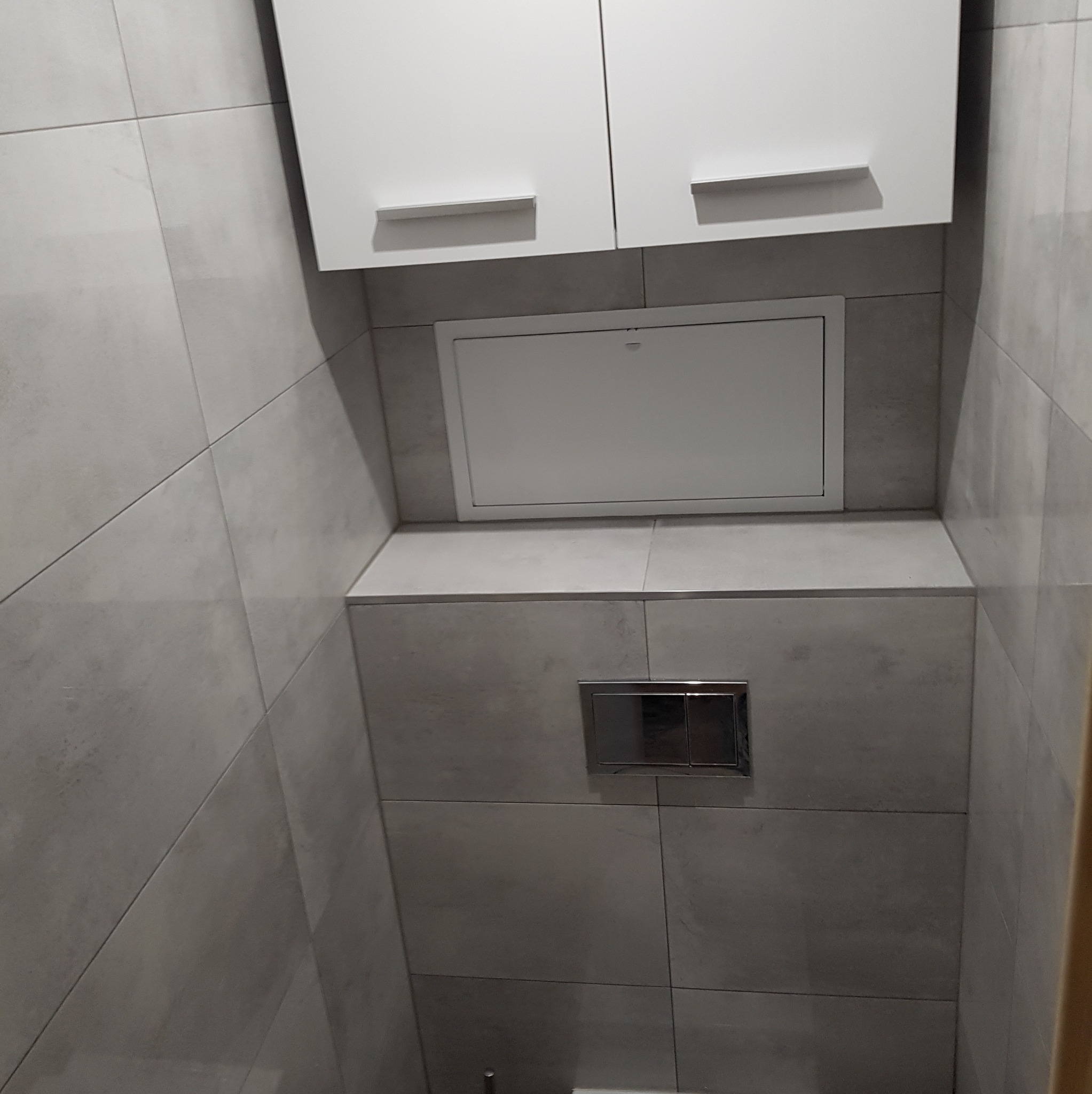 Szara łazienka z zabudową WC, szafką nad stelażem i ukrytym włazem rewizyjnym. Widoczny chromowany przycisk spłuczki.