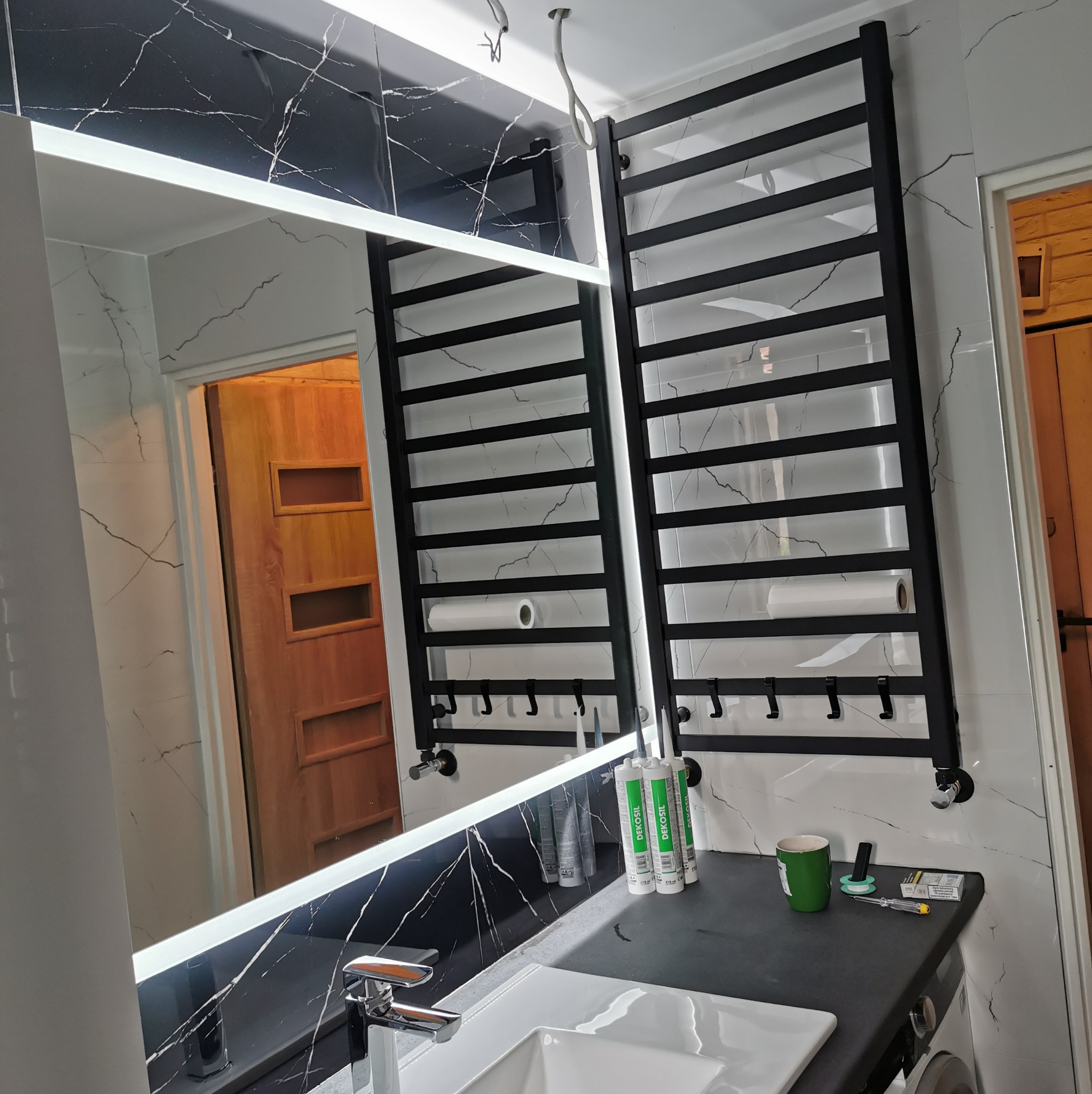 Łazienka w trakcie wykańczania: czarny grzejnik drabinkowy z ręcznikami, lustro z podświetleniem LED, białe płytki z czarnymi żyłkami, widoczne silikony i narzędzia na blacie.