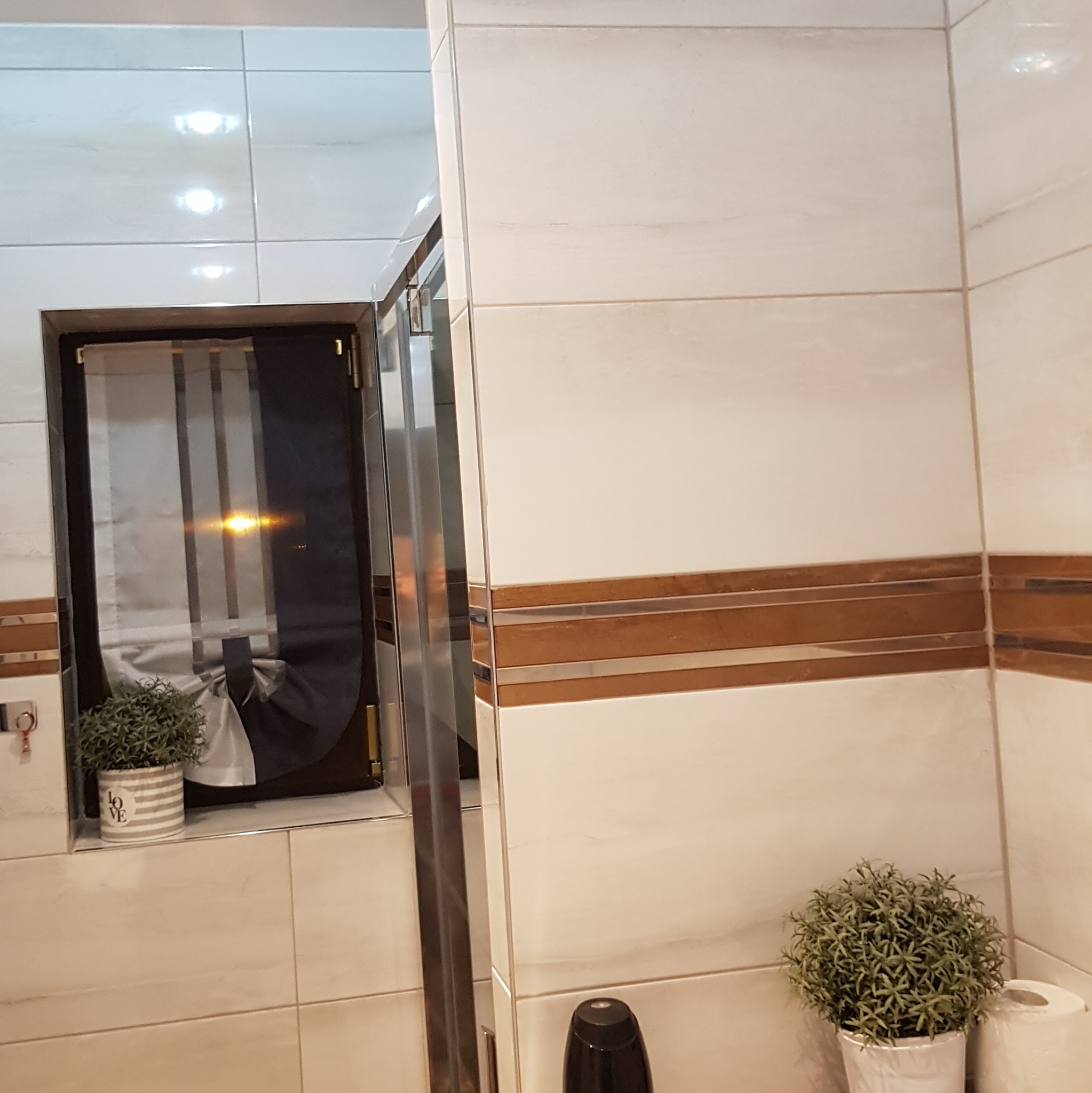 Wykończona łazienka z beżowymi płytkami, dekoracyjnym pasem, czarną roletą w oknie i dwoma sztucznymi roślinami w doniczkach.