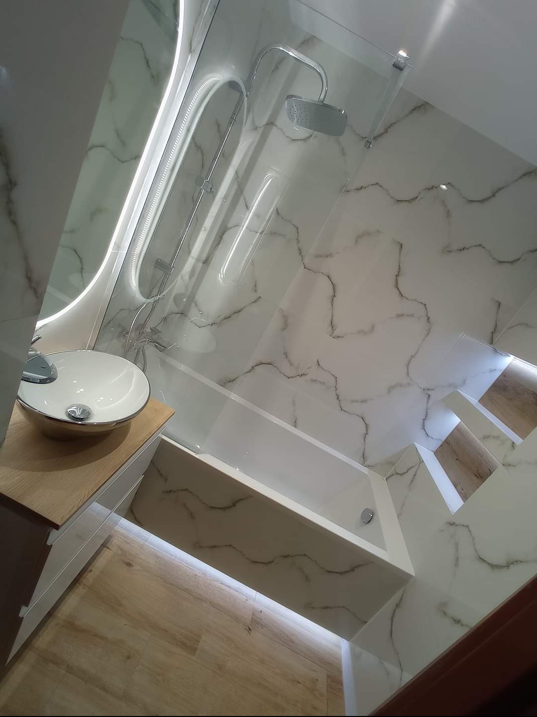 Nowoczesna łazienka z marmurowymi płytkami, podświetlaną wanną i lustrem z oświetleniem LED, umywalka nablatowa na drewnianej szafce, widok z góry.