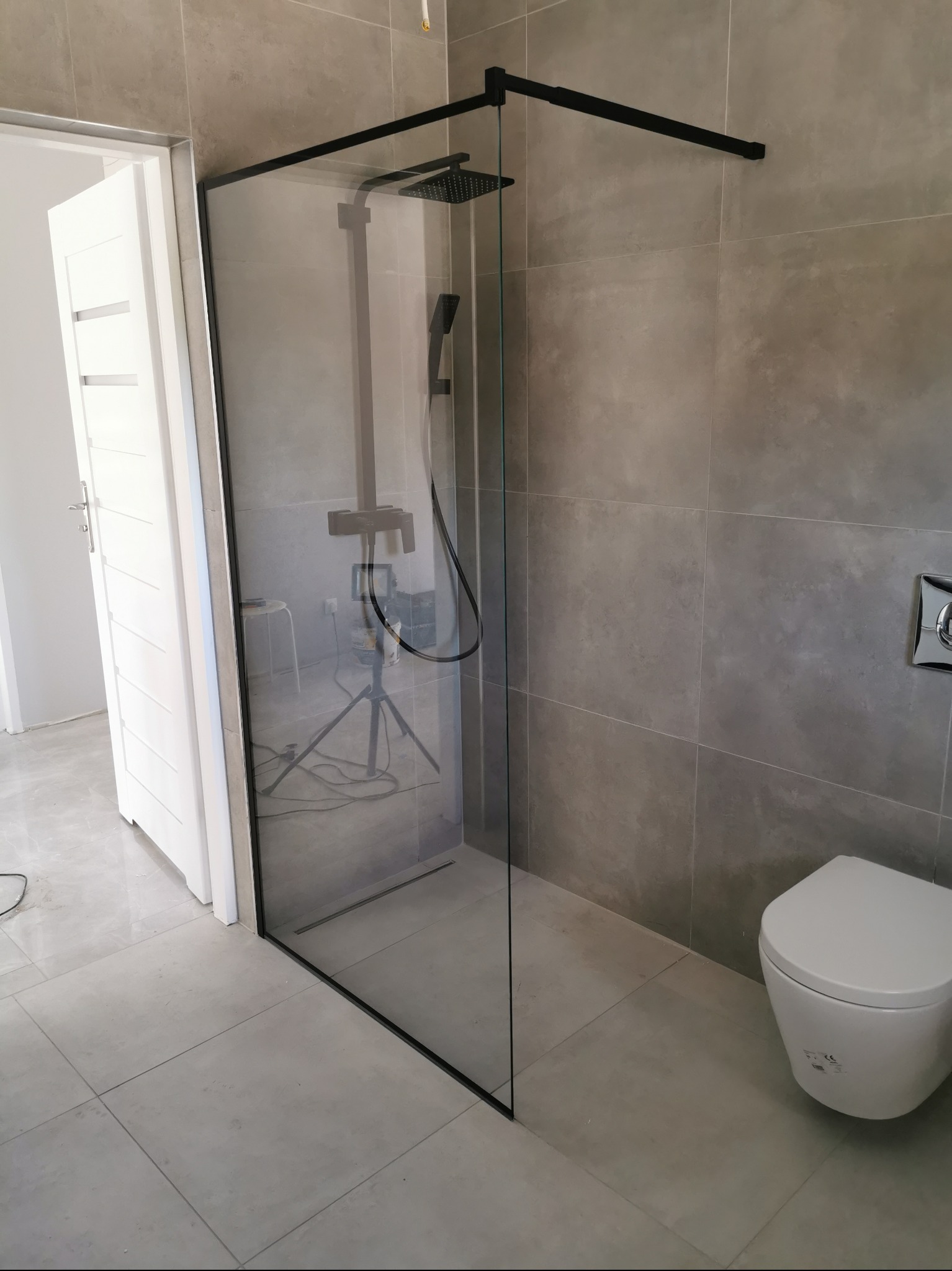 Nowoczesna kabina prysznicowa typu walk-in z czarnymi profilami, zamontowana w łazience z szarymi płytkami, widoczny biały sedes i częściowo otwarte białe drzwi, wewnątrz kabiny widoczny statyw...