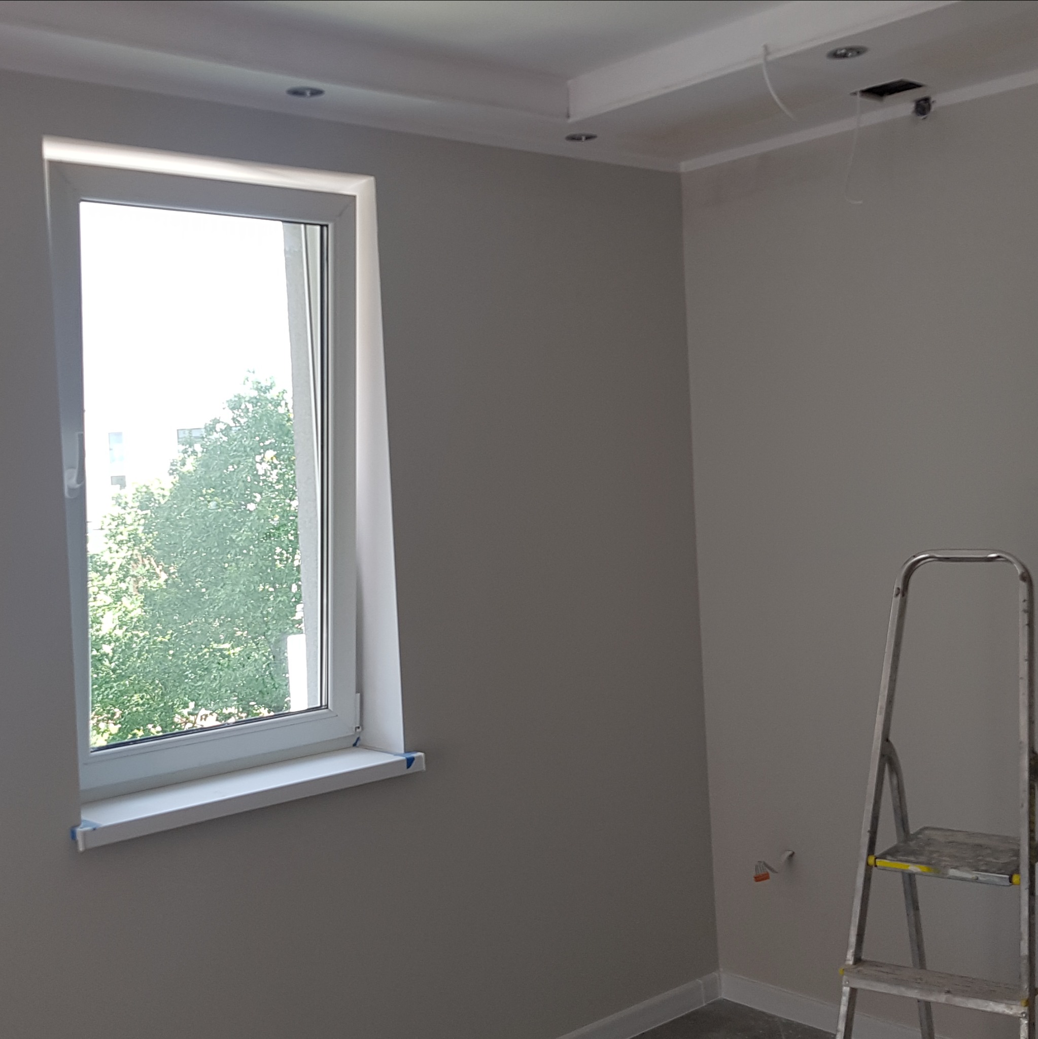 Wnętrze pokoju w trakcie remontu: pomalowane szare ściany, białe okno z widokiem na drzewo, aluminiowa drabina i przygotowane otwory na oświetlenie w suficie.