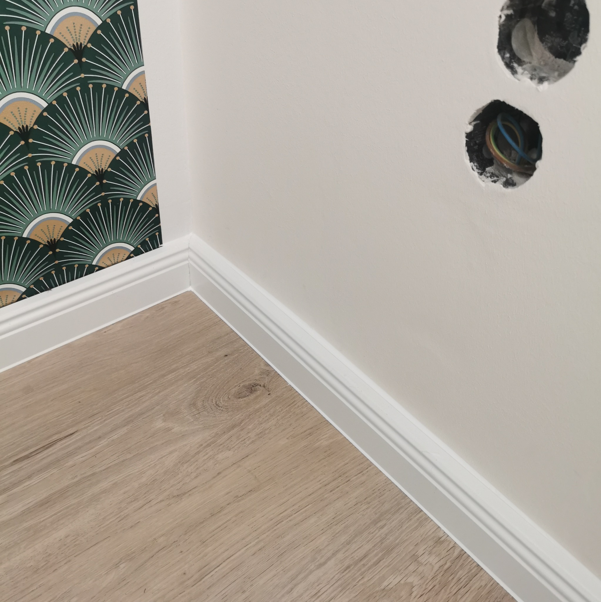 Fragment wykończonego wnętrza: panelowa podłoga, białe listwy przypodłogowe, ściana z wzorzystą tapetą w stylu art déco, gładka ściana z dwoma otworami na gniazdka elektryczne z widocznymi przewodami.