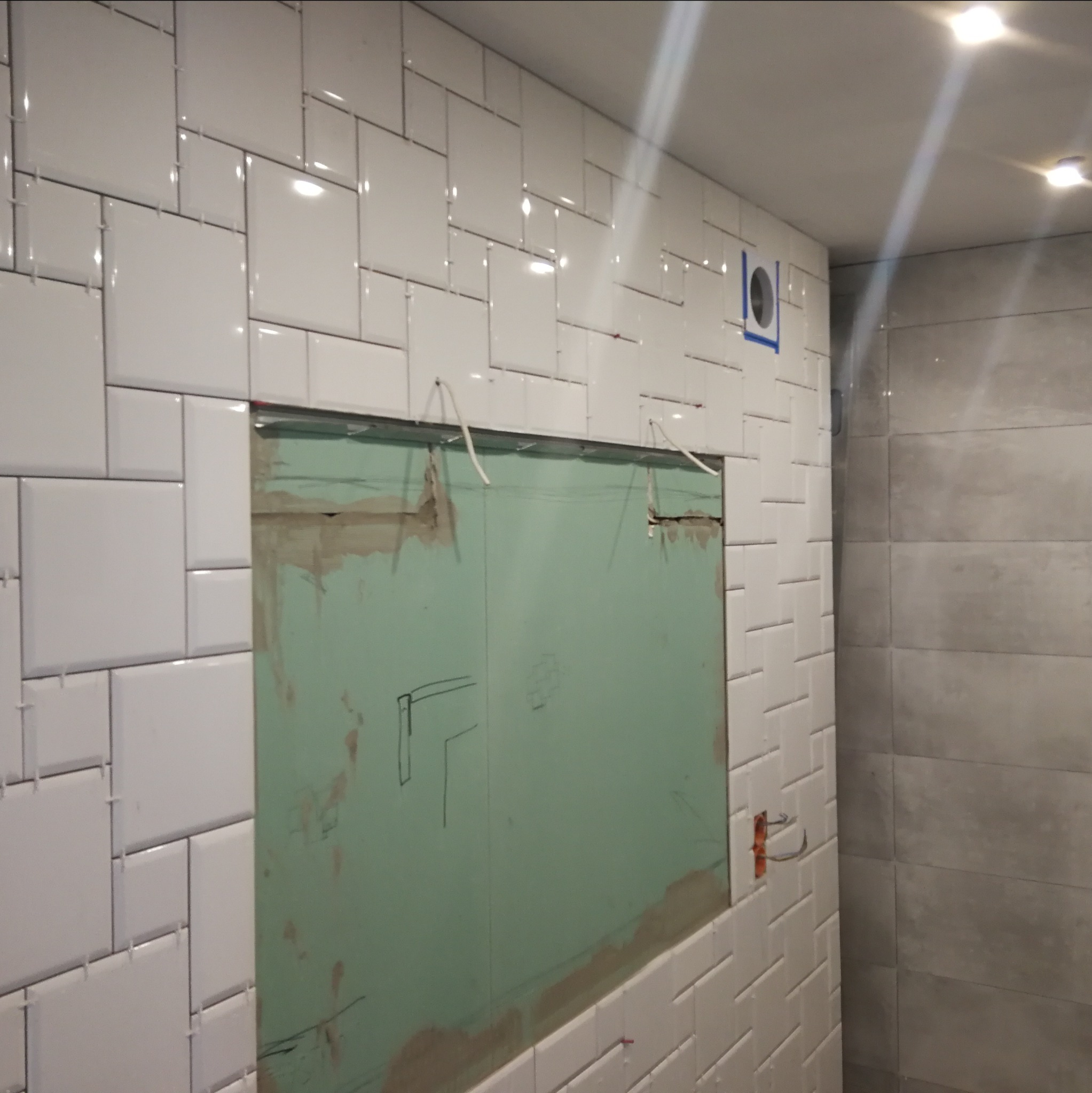 Ściana w trakcie układania białych, prostokątnych płytek; widoczna wnęka z zieloną płytą kartonowo-gipsową z naniesionymi szkicami, wystające kable elektryczne i otwór wentylacyjny zabezpieczony...