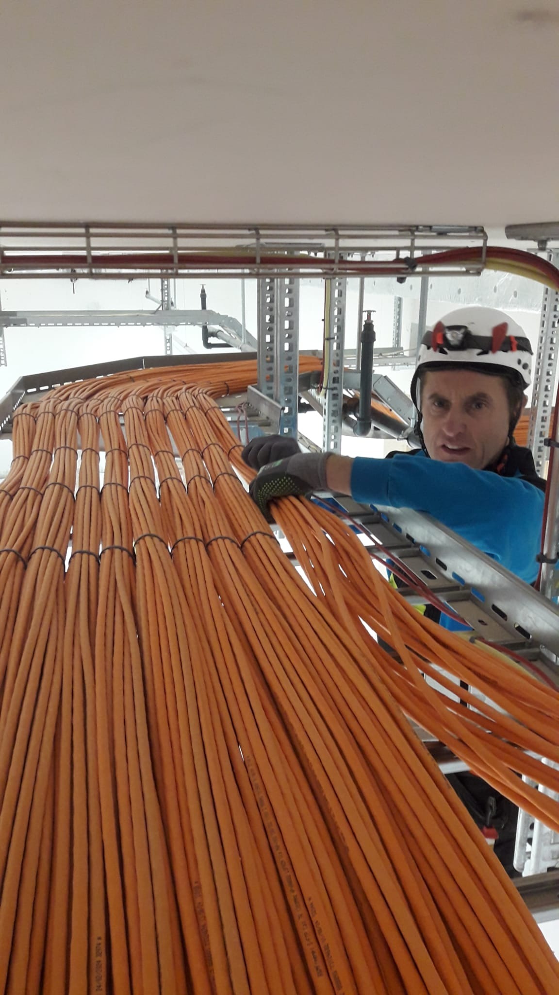 Elektryk w kasku z latarką układa pomarańczowe kable w korycie kablowym, widoczne metalowe konstrukcje nośne.