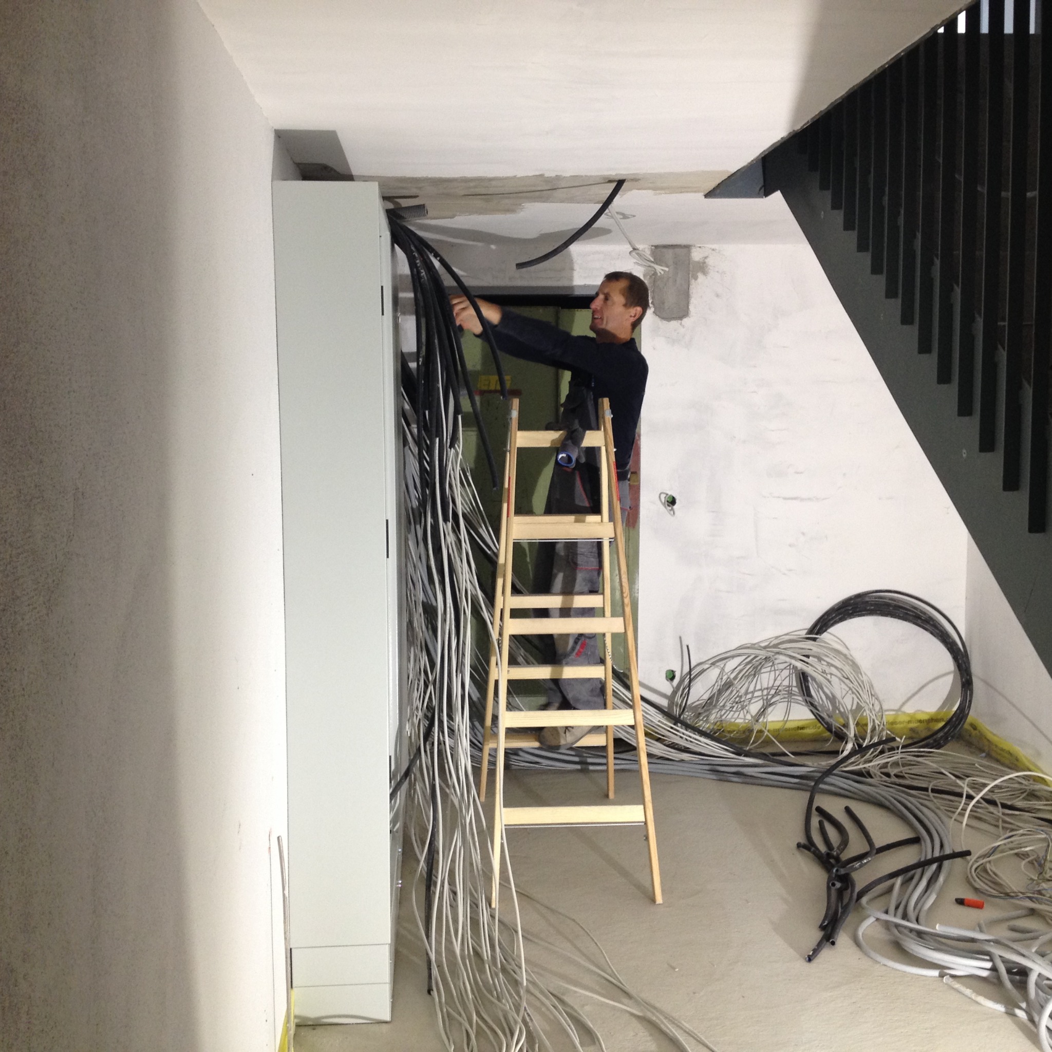 Elektryk na drewnianej drabinie, układa wiązki przewodów w nowym budynku w pobliżu białej szafy sterowniczej; w tle widoczne schody i surowy stan wykończenia wnętrza.