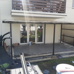 AR Adam Ozimek - Zadaszenie tarasu z poliwęglanu w trakcie montażu, widoczne narzędzia i materiały budowlane, w tle balkon z drewnianą balustradą i pies w oknie.