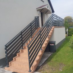 AR Adam Ozimek - Zewnętrzne schody z terakoty prowadzące do domu, zabezpieczone nowoczesną, ciemnoszarą metalową balustradą o poziomym układzie szczebli, z widocznymi koszami na śmieci po jednej stronie...