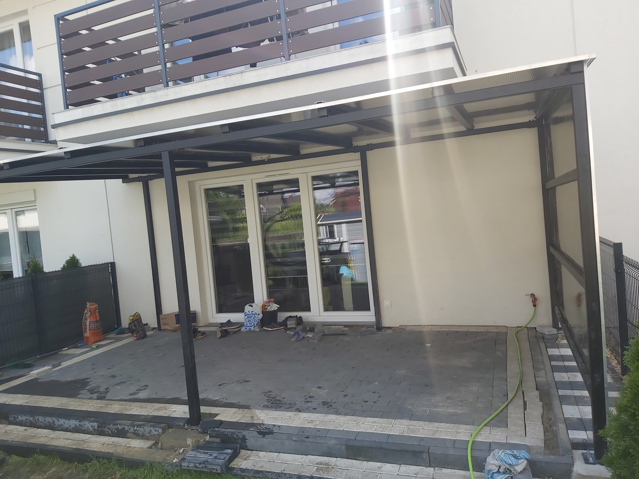 Zadaszenie tarasu z metalową konstrukcją i przezroczystym dachem poliwęglanowym, widoczne narzędzia i materiały budowlane na kostce brukowej.