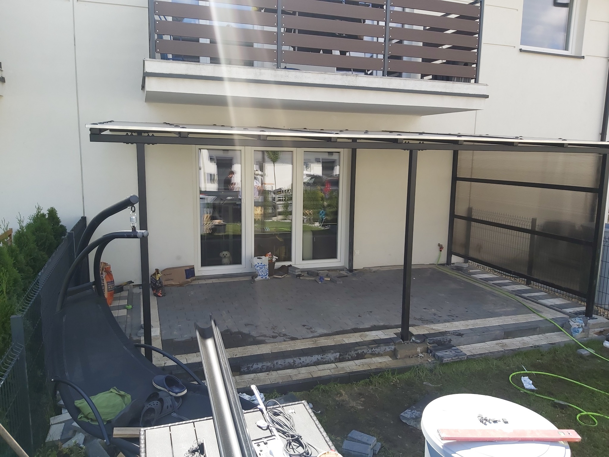 Zadaszenie tarasu z poliwęglanu w trakcie montażu, widoczne narzędzia i materiały budowlane, w tle balkon z drewnianą balustradą i pies w oknie.