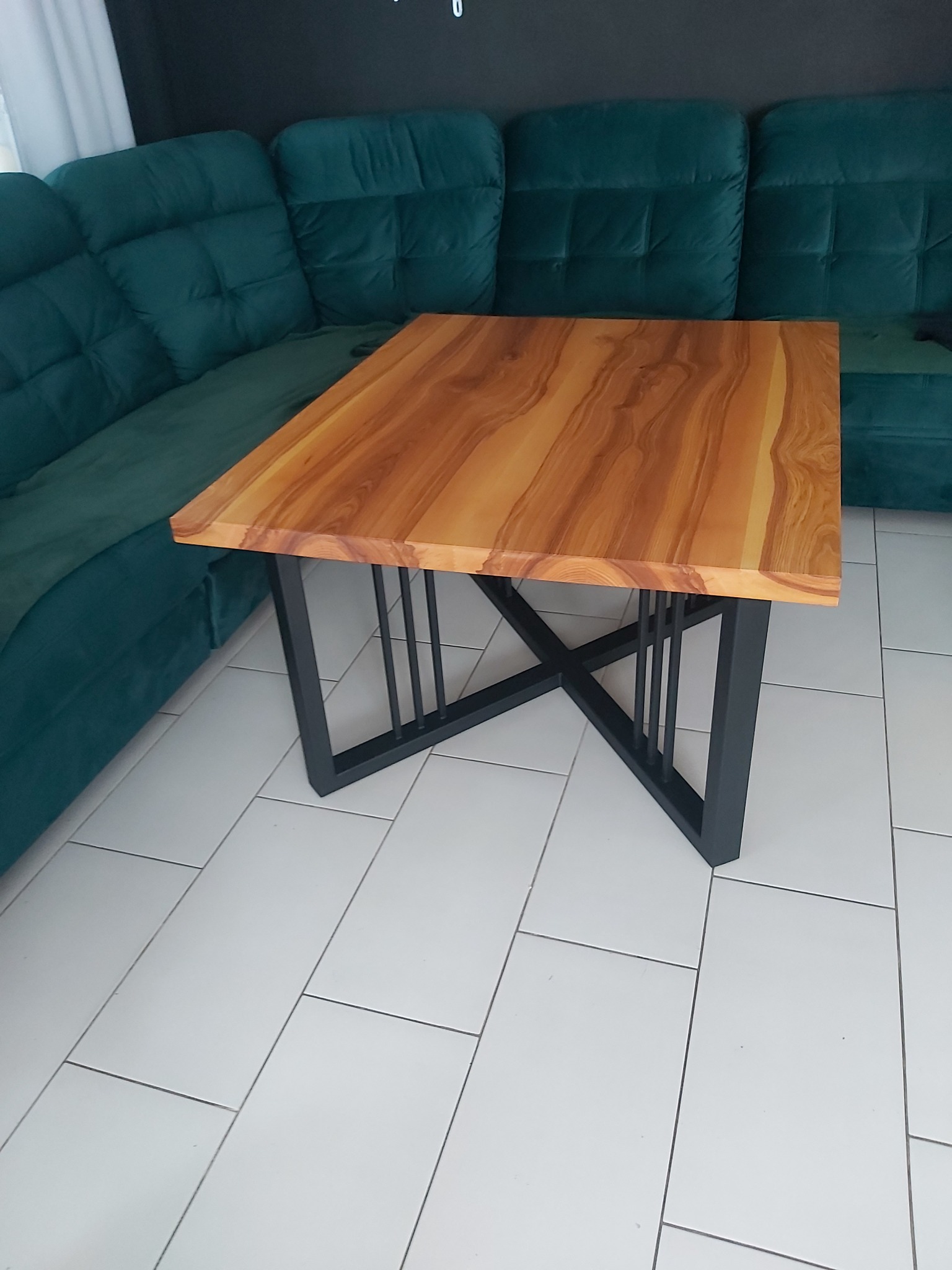 Stolik kawowy z blatem z jasnego drewna o wyraźnym usłojeniu i czarną, metalową podstawą z pionowymi prętami, na tle turkusowej, pikowanej sofy i białej podłogi z płytek.