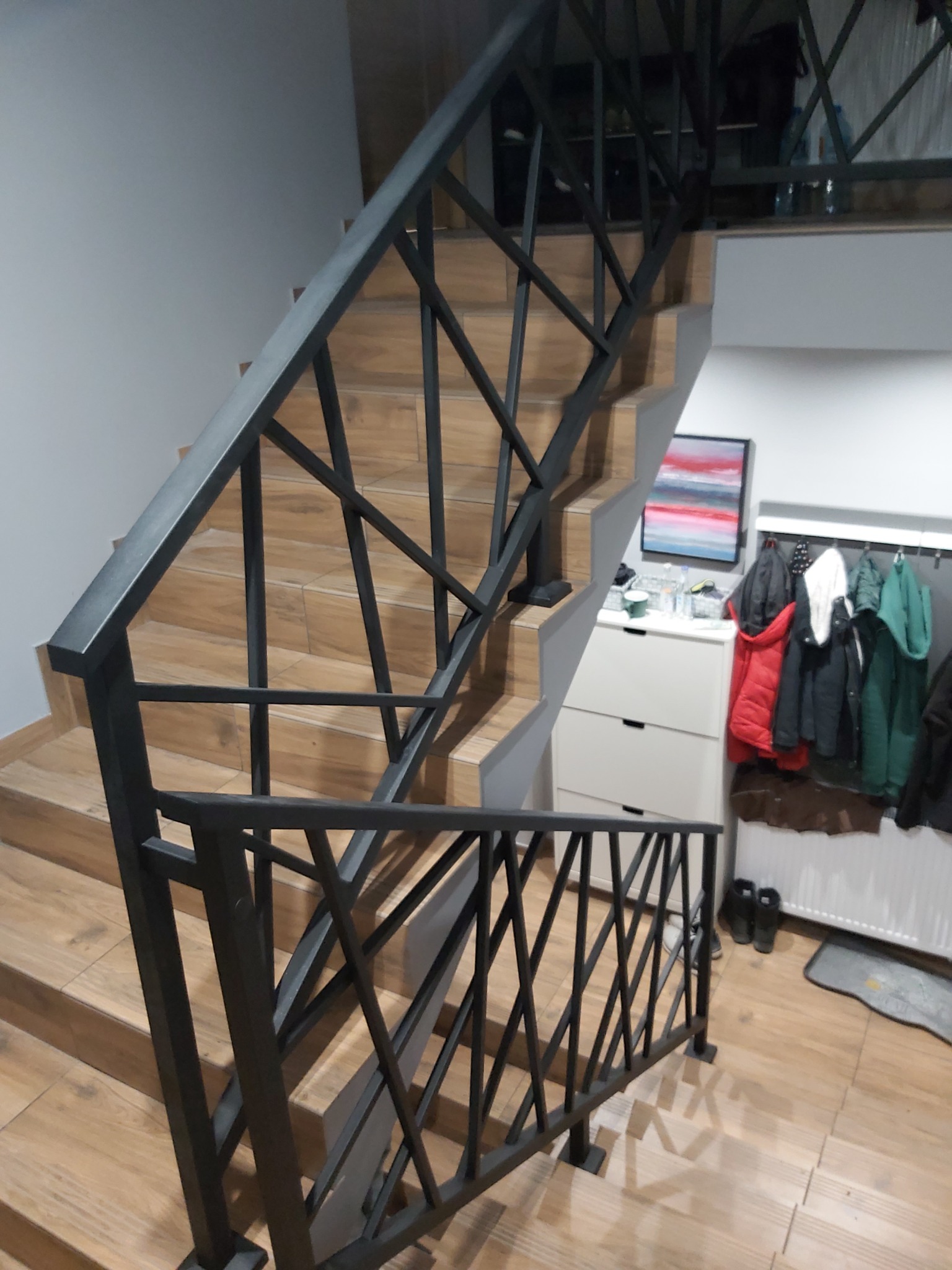 Nowoczesne schody z drewnopodobnymi płytkami i czarną, geometryczną balustradą metalową, widok z dołu na górę.