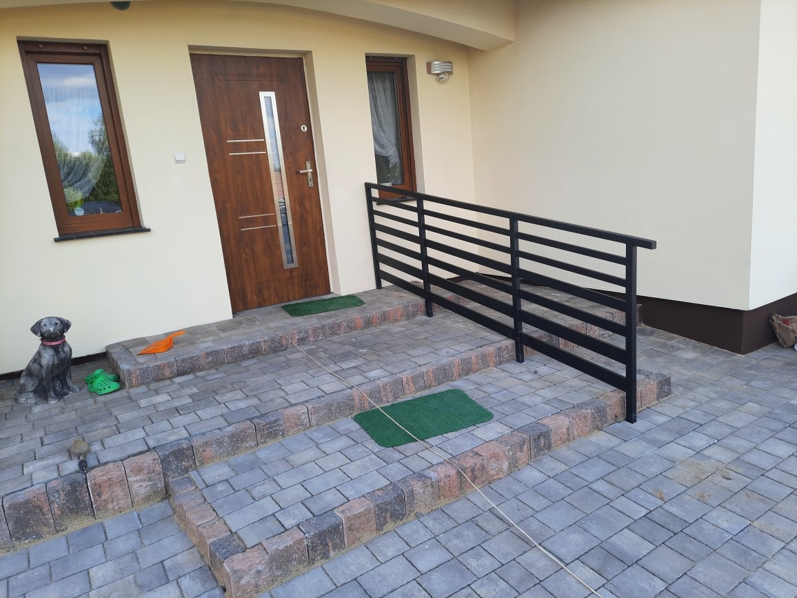 Czarna, pozioma balustrada przed wejściem do domu, z trzema stopniami wyłożonymi kostką brukową, widok na wejście z brązowymi drzwiami i małym oknem, obok drzwi dekoracja w postaci figurki psa...