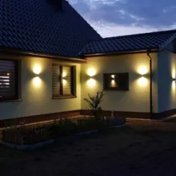 Elewacja domu jednorodzinnego oświetlona symetrycznie rozmieszczonymi lampami ściennymi, widoczne okna z żaluzjami, ogródek przed domem, wieczorne niebo.