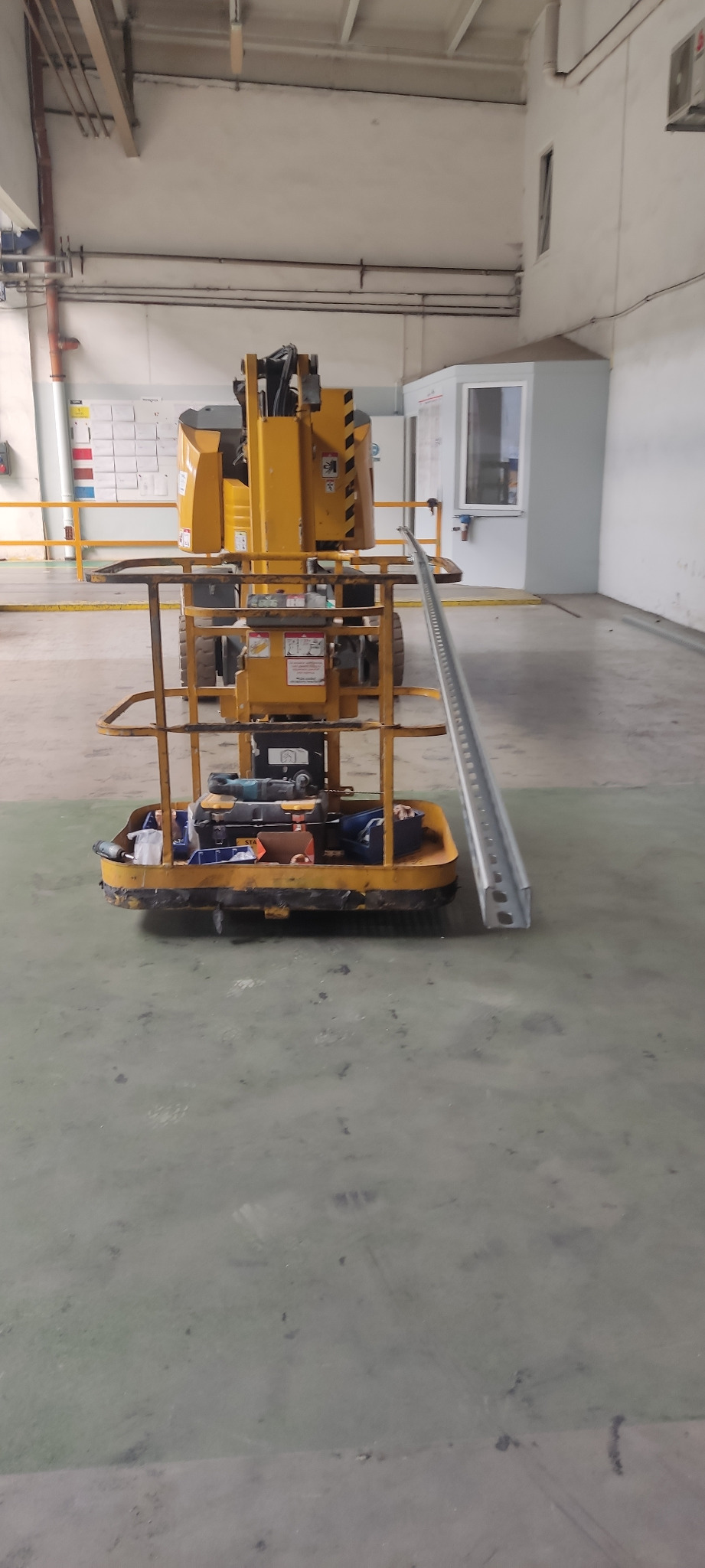 Żółty podnośnik koszowy z narzędziami w koszu stoi w hali przemysłowej obok metalowego profilu.