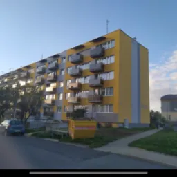 Wielopiętrowy budynek mieszkalny z balkonami, częściowo odnowiony, z żółtą i szarą elewacją, widoczne rusztowania, kontener na odpady budowlane i zaparkowane samochody.